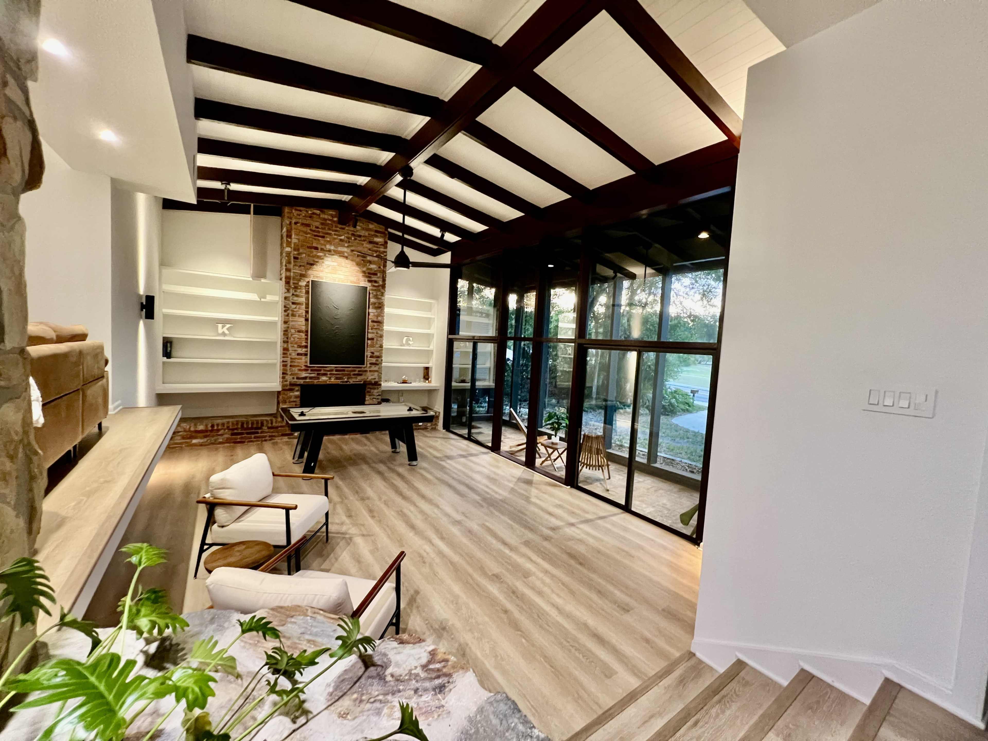 A spacious living area with high, beamed ceilings, large glass windows, and a modern design featuring a pool table and a staircase.