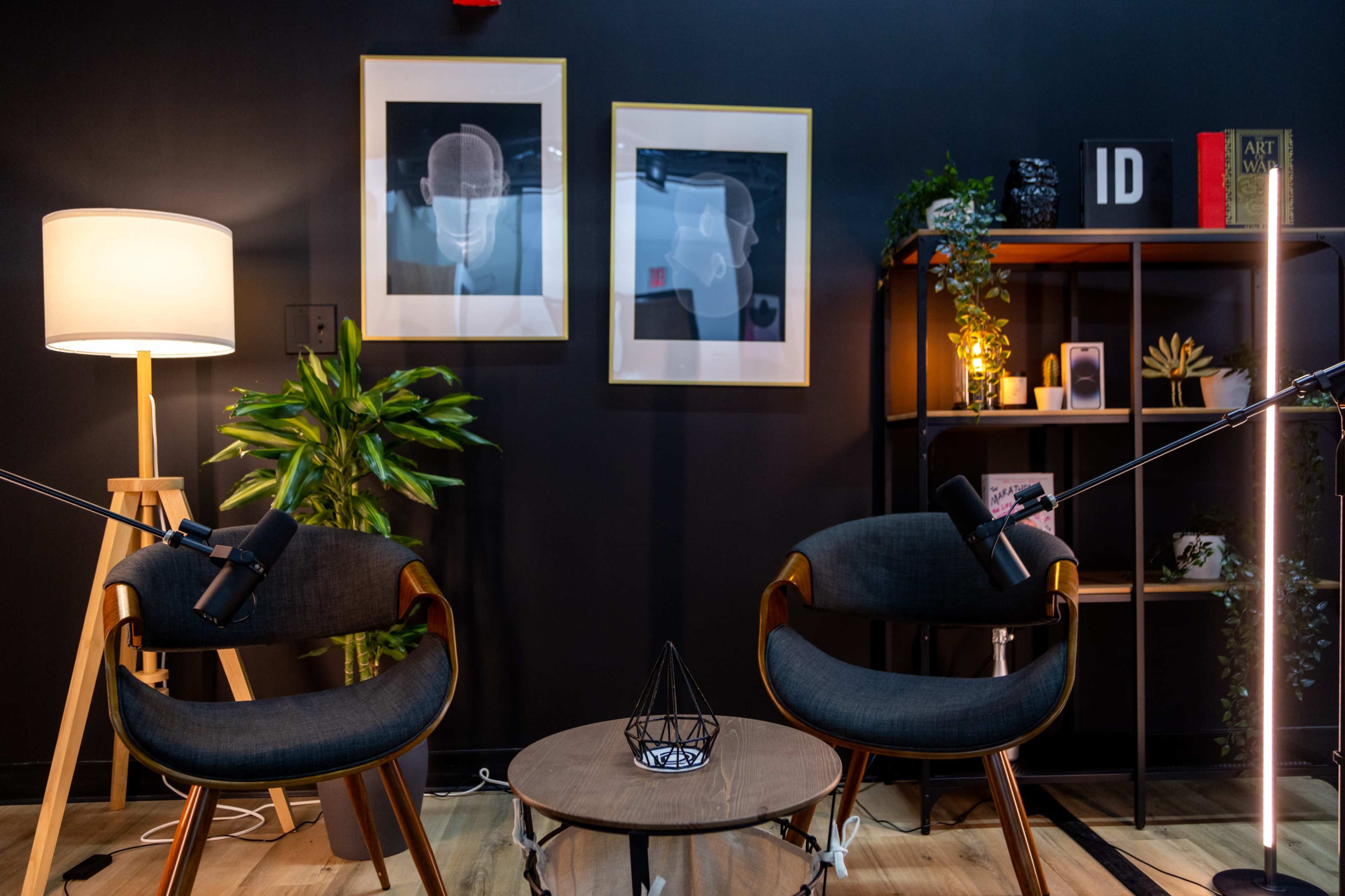 A modern podcast studio features two chairs, a round table, and decorative elements against a dark wall adorned with framed artwork.