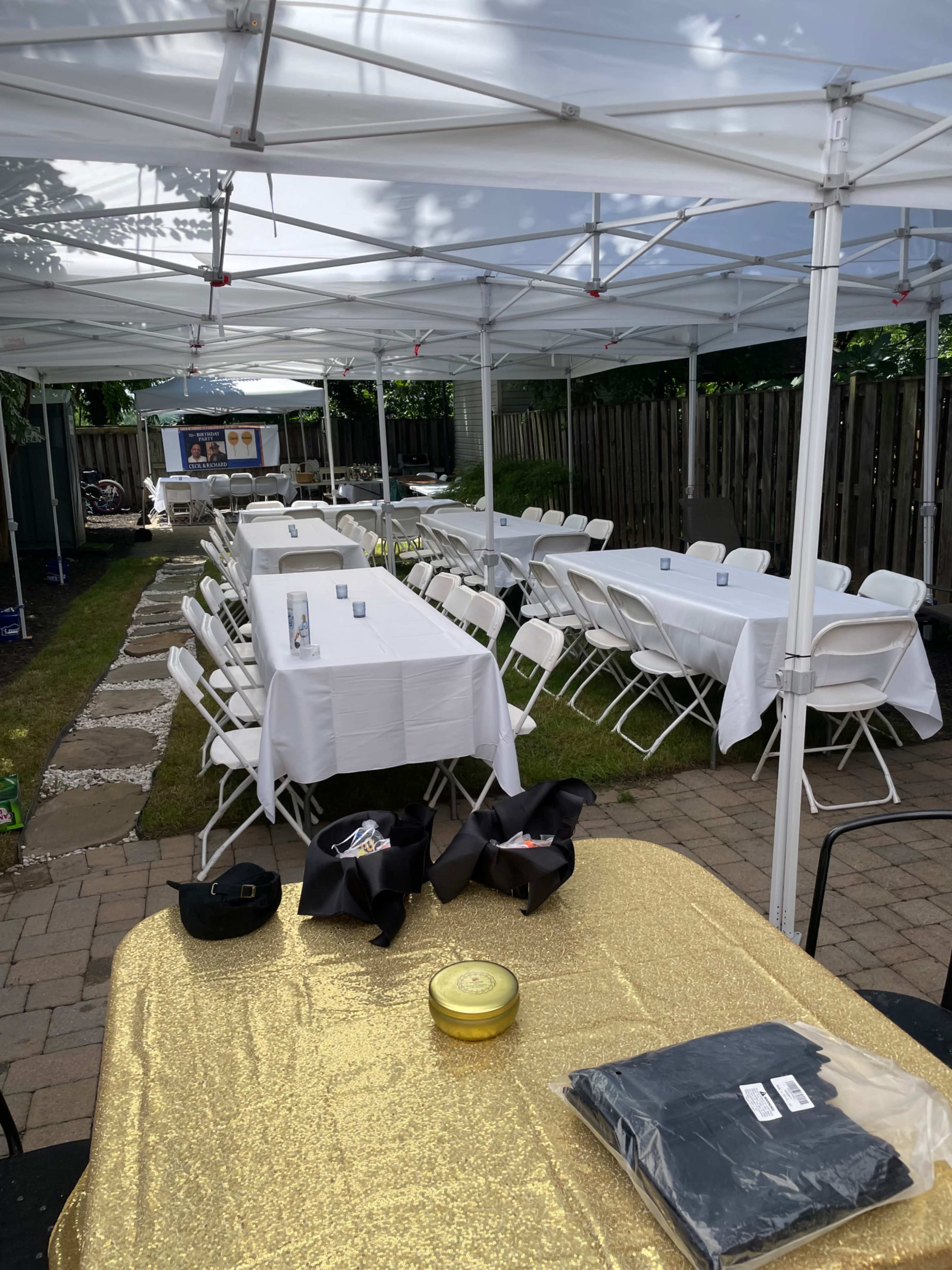 A long outdoor setup features rows of white tables and chairs under canopies, with a gold tablecloth in the foreground.