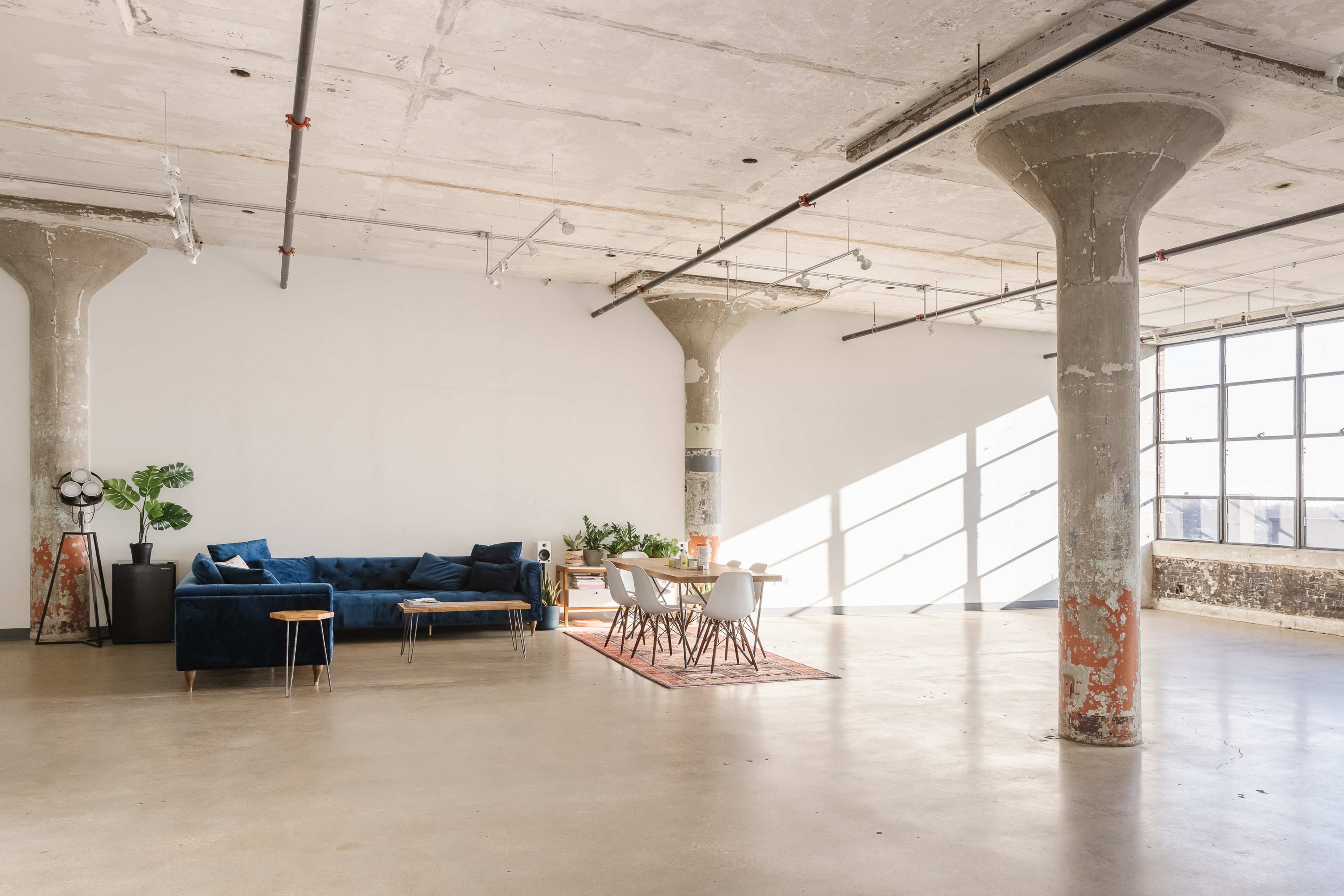 The image shows a spacious, modern interior with a blue sofa, a dining table, and large windows allowing natural light to fill the room.