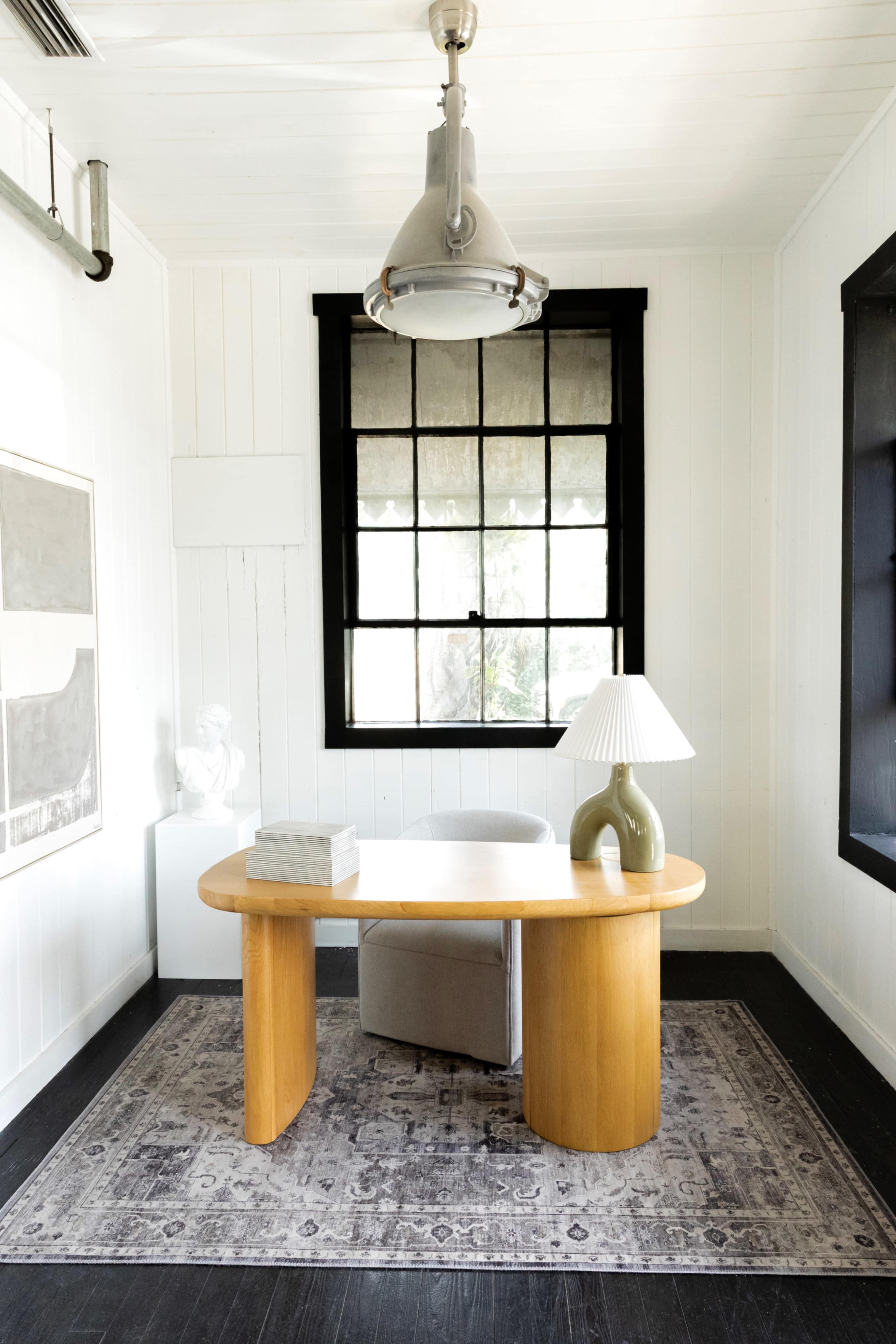 A wooden desk with a curved edge is positioned in a well-lit room, accompanied by a chair, a lamp, and decorative items on a patterned rug.