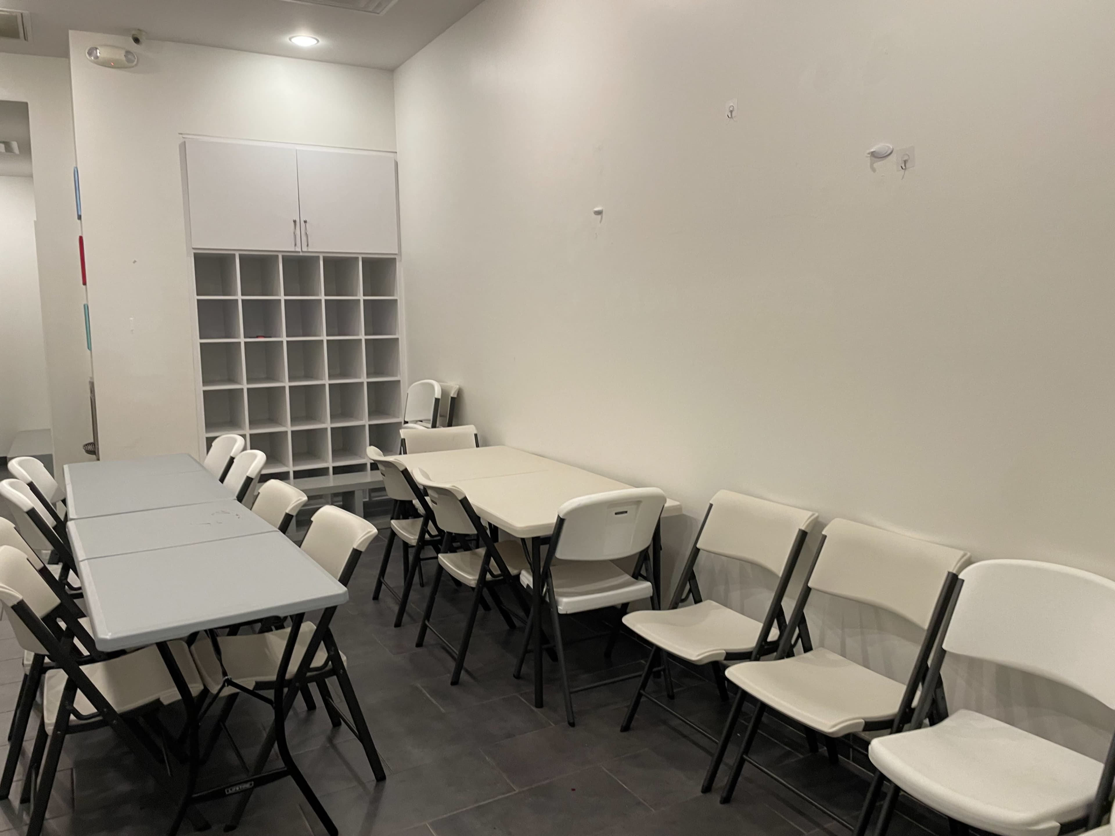 The image shows a room with several folding tables and chairs arranged in a simple layout, along with a storage cabinet against the wall.
