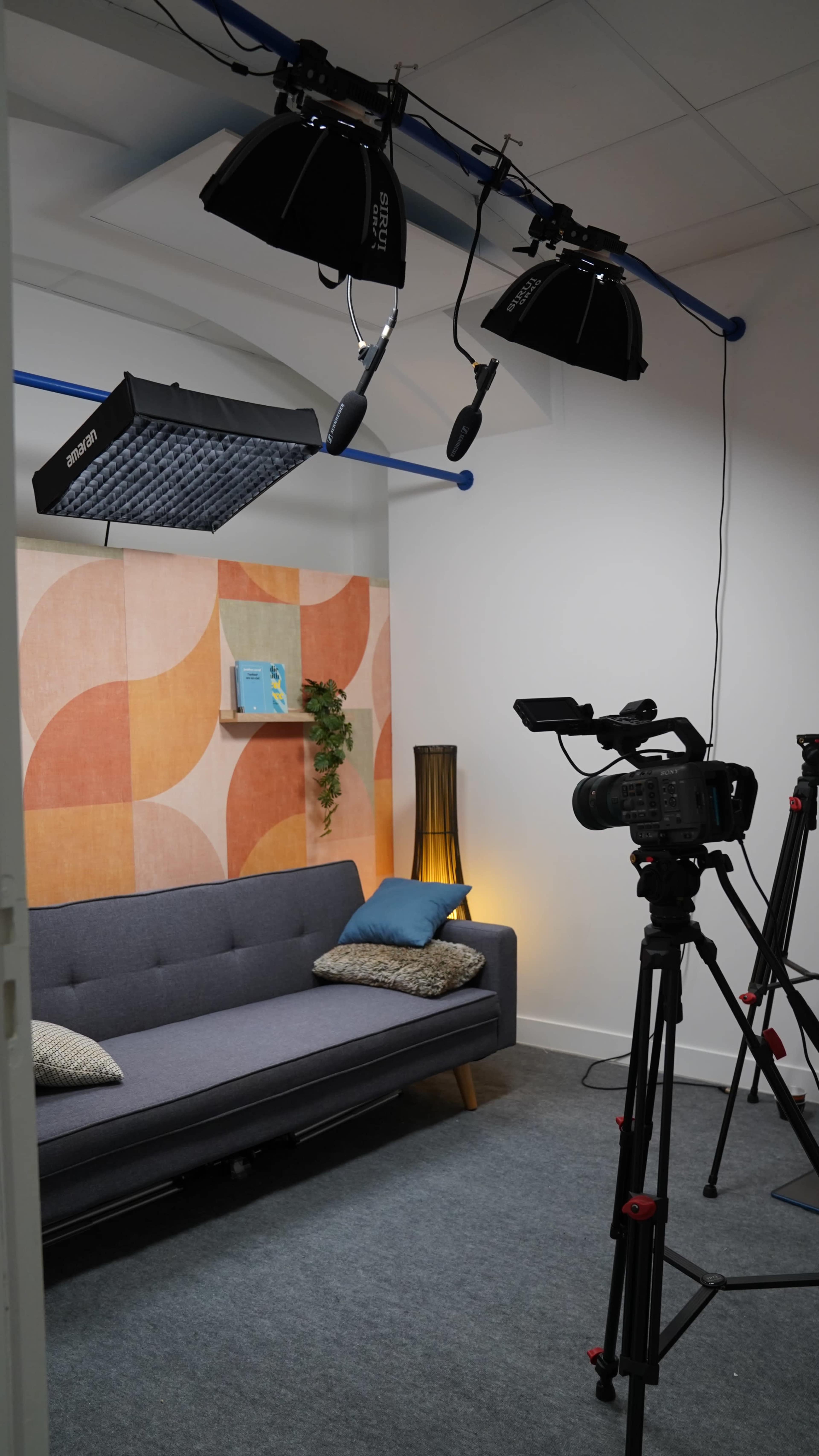 The image shows a well-lit studio setup featuring a gray couch, a wall with colorful geometric patterns, and camera equipment positioned for filming.