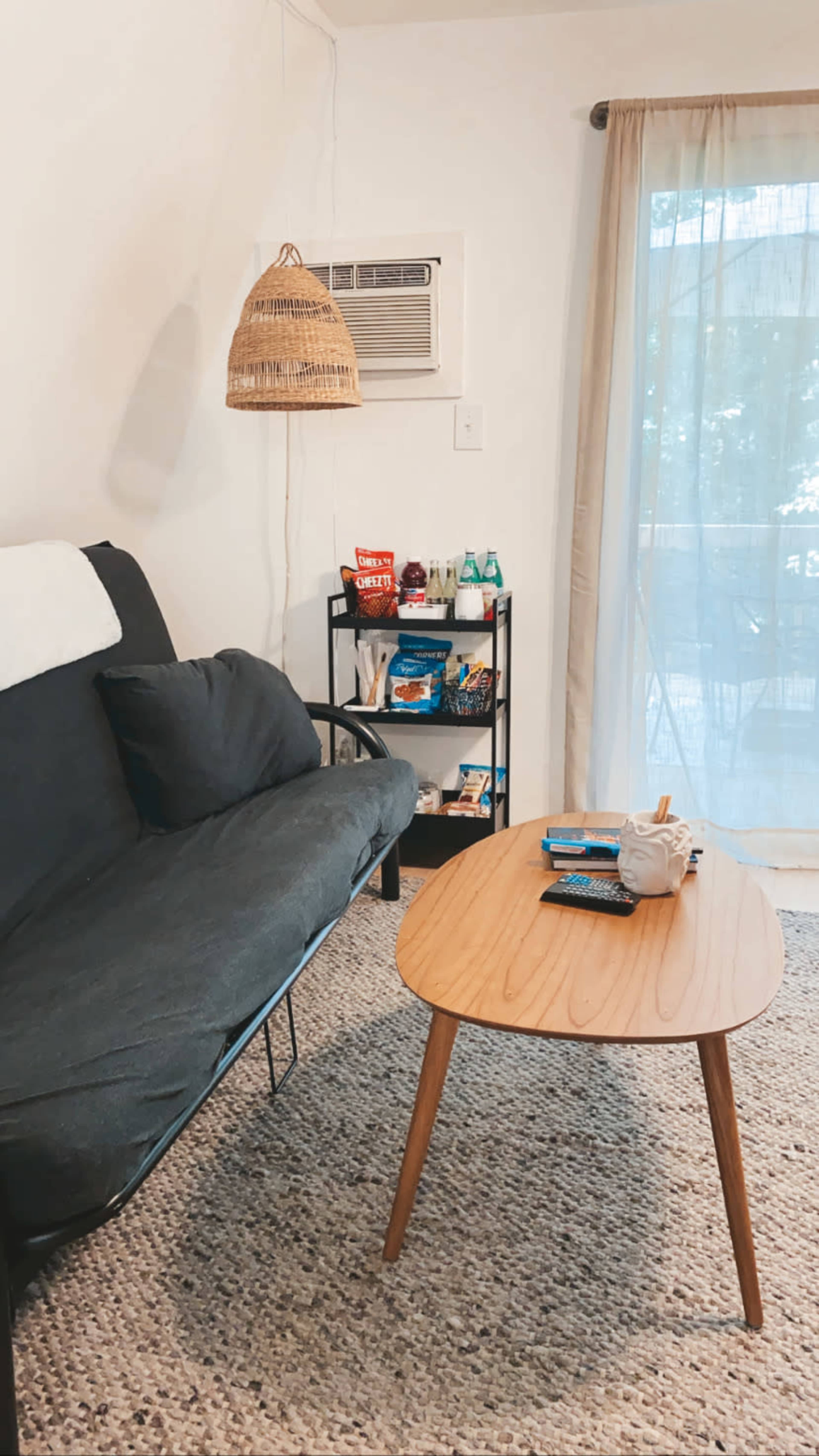 The image shows a cozy living room with a dark sofa, a wooden coffee table, and a small shelving unit filled with various items, illuminated by natural light through a sheer curtain.