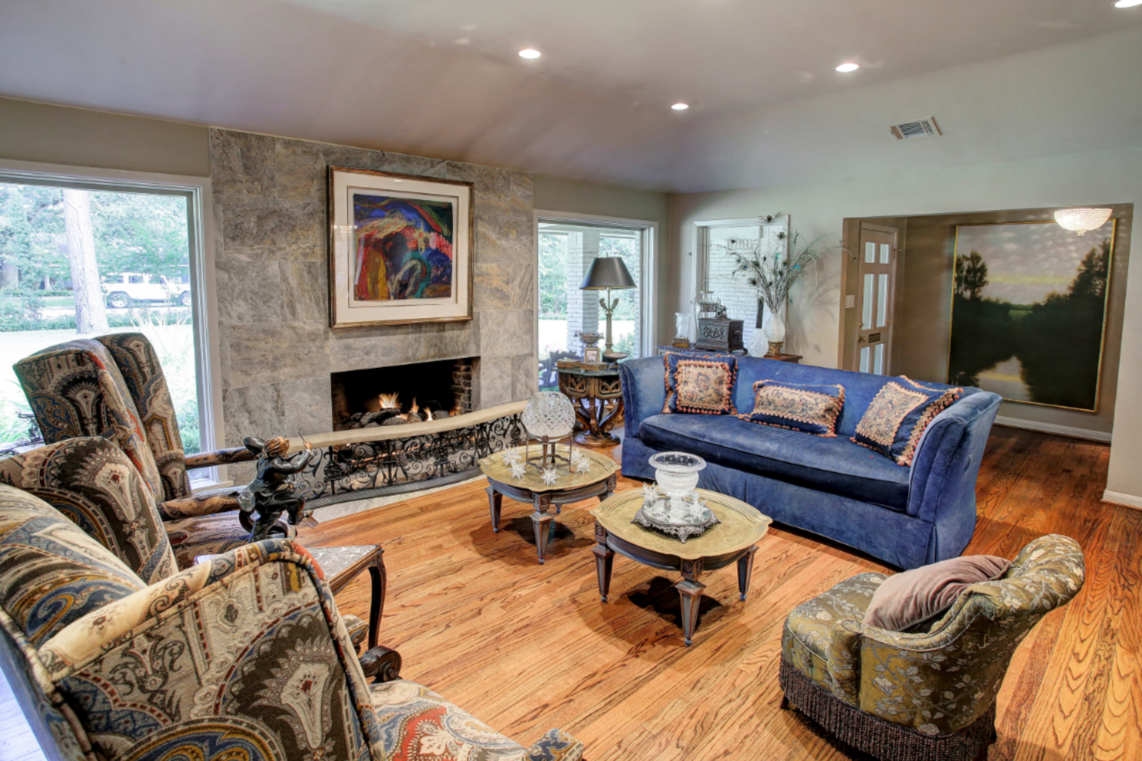 The image depicts a living room with a blue sofa, patterned armchairs, a stone fireplace, and large windows allowing natural light to enter.