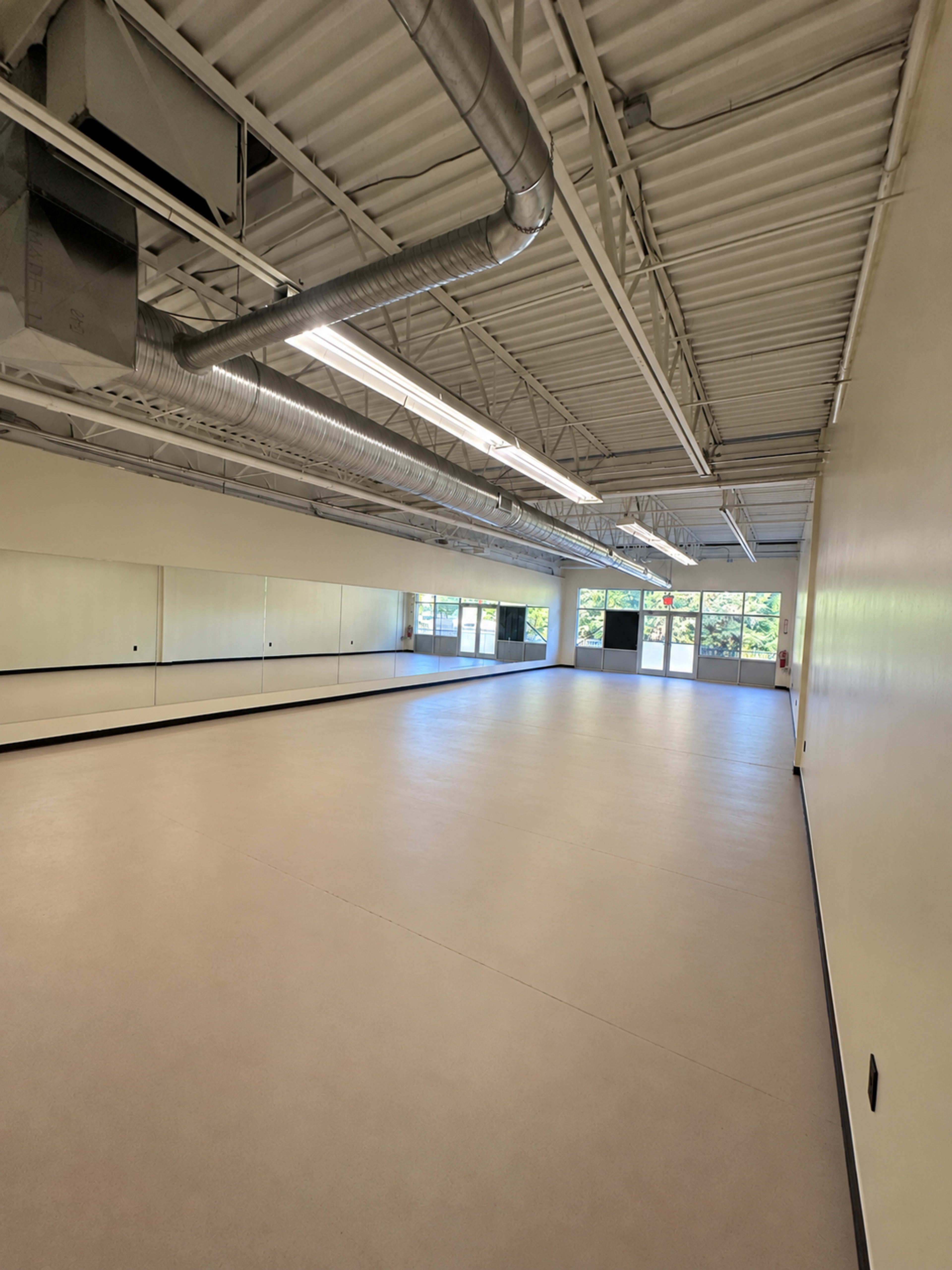 The image shows a spacious, empty room with mirrored walls, bright overhead lighting, and large windows on one side.