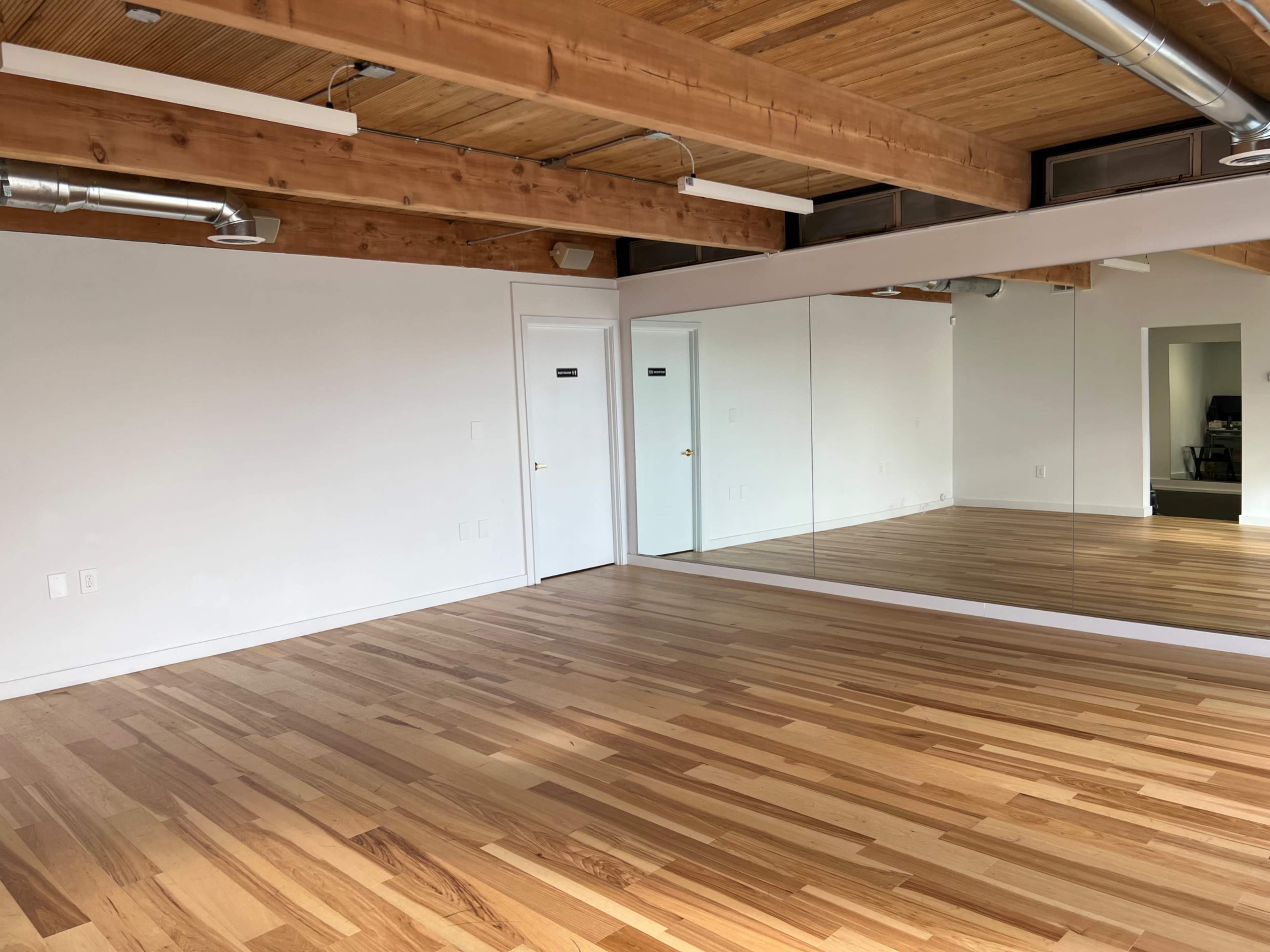 A spacious room with a hardwood floor, exposed wooden beams, and a large mirror on one wall.