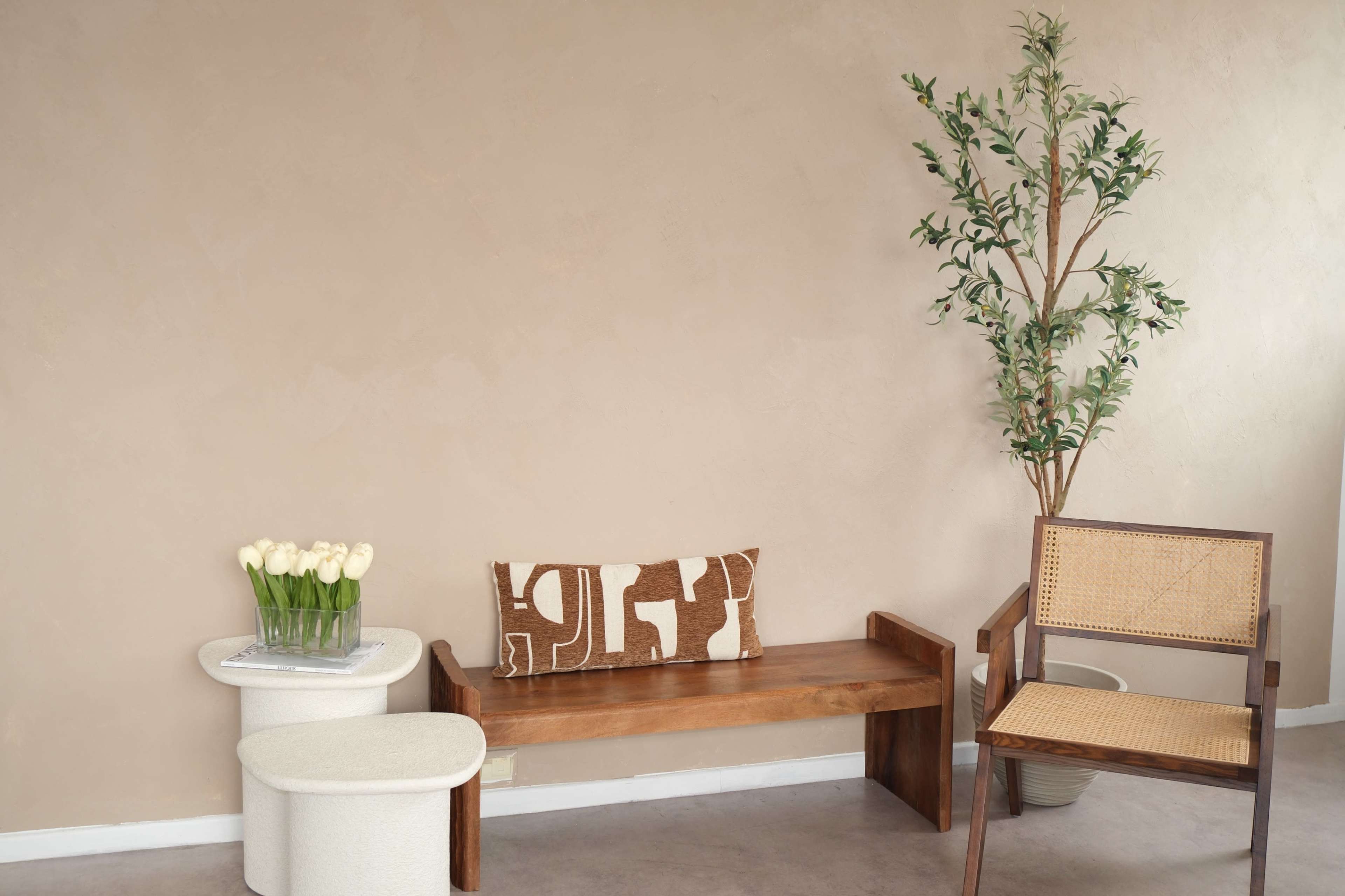 The scene features a minimalist room with a wooden bench, a round side table, a chair, and a potted plant, all against a beige wall.