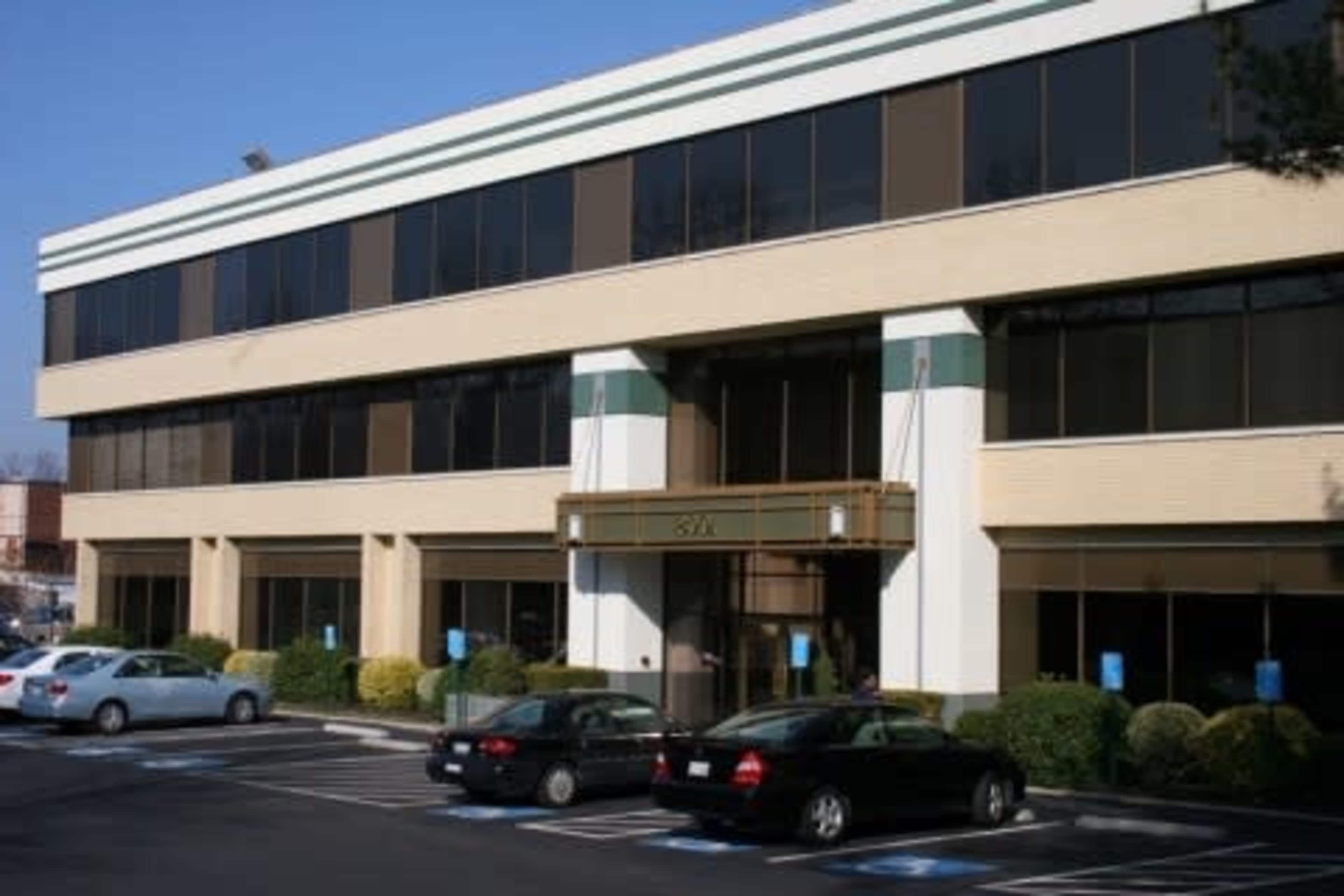 A three-story office building with large windows and a central entrance, surrounded by a parking lot with several cars.
