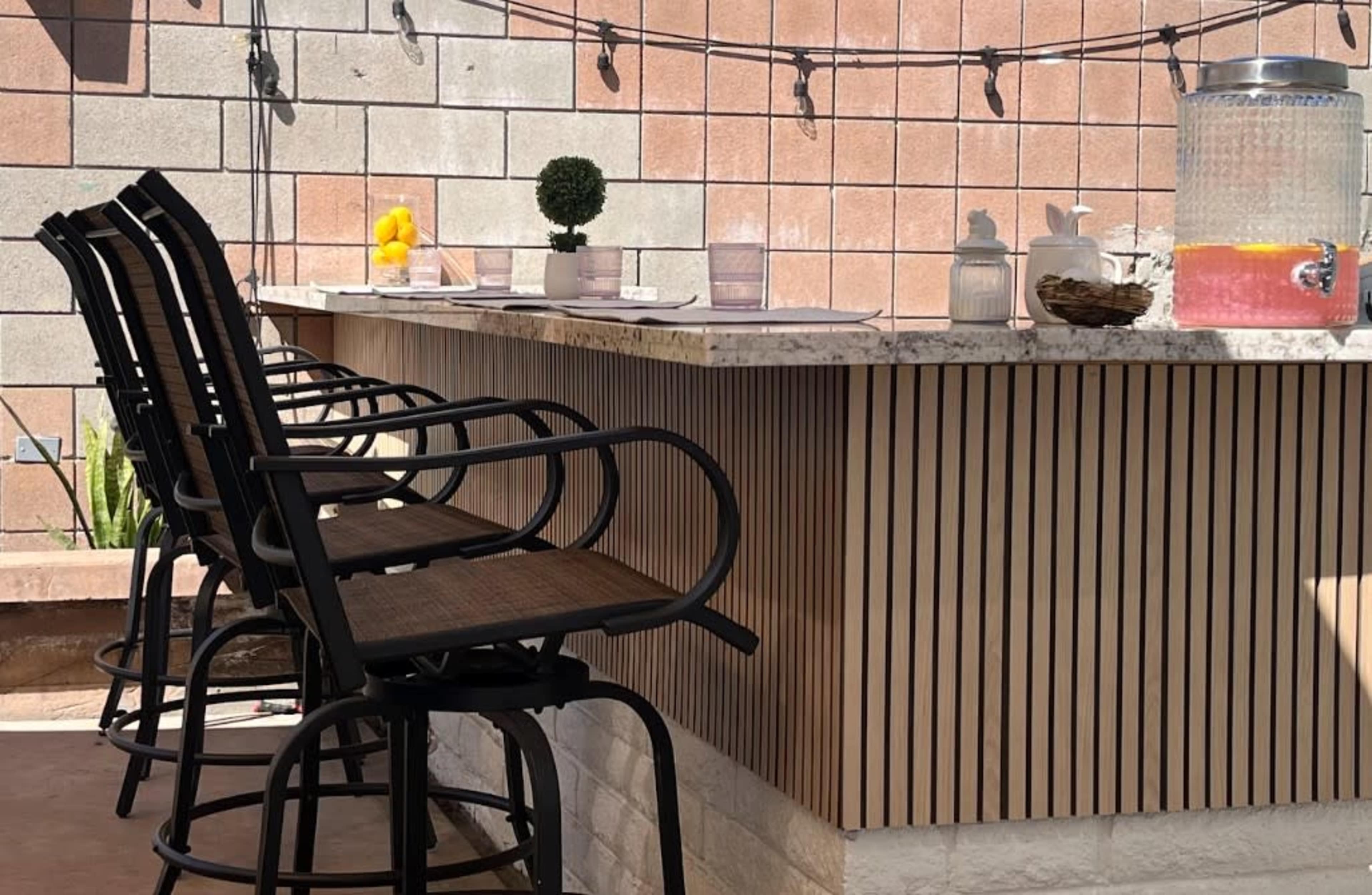 The image shows a bar area with black, tall stools lined up in front of a marble counter, complemented by a drink dispenser and decorative items against a textured wall.