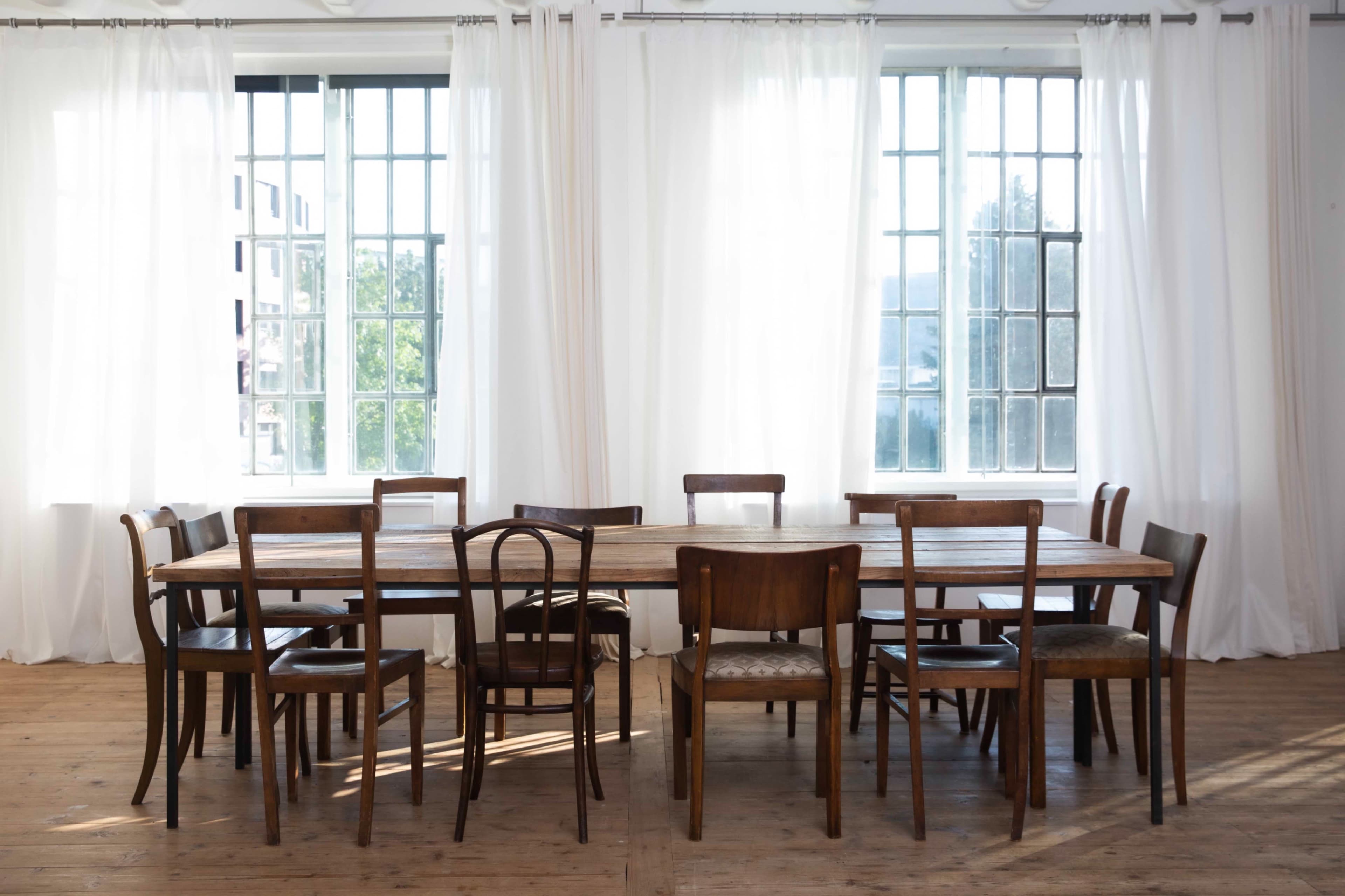 A long wooden table surrounded by chairs is positioned in a bright room with large windows and sheer white curtains.