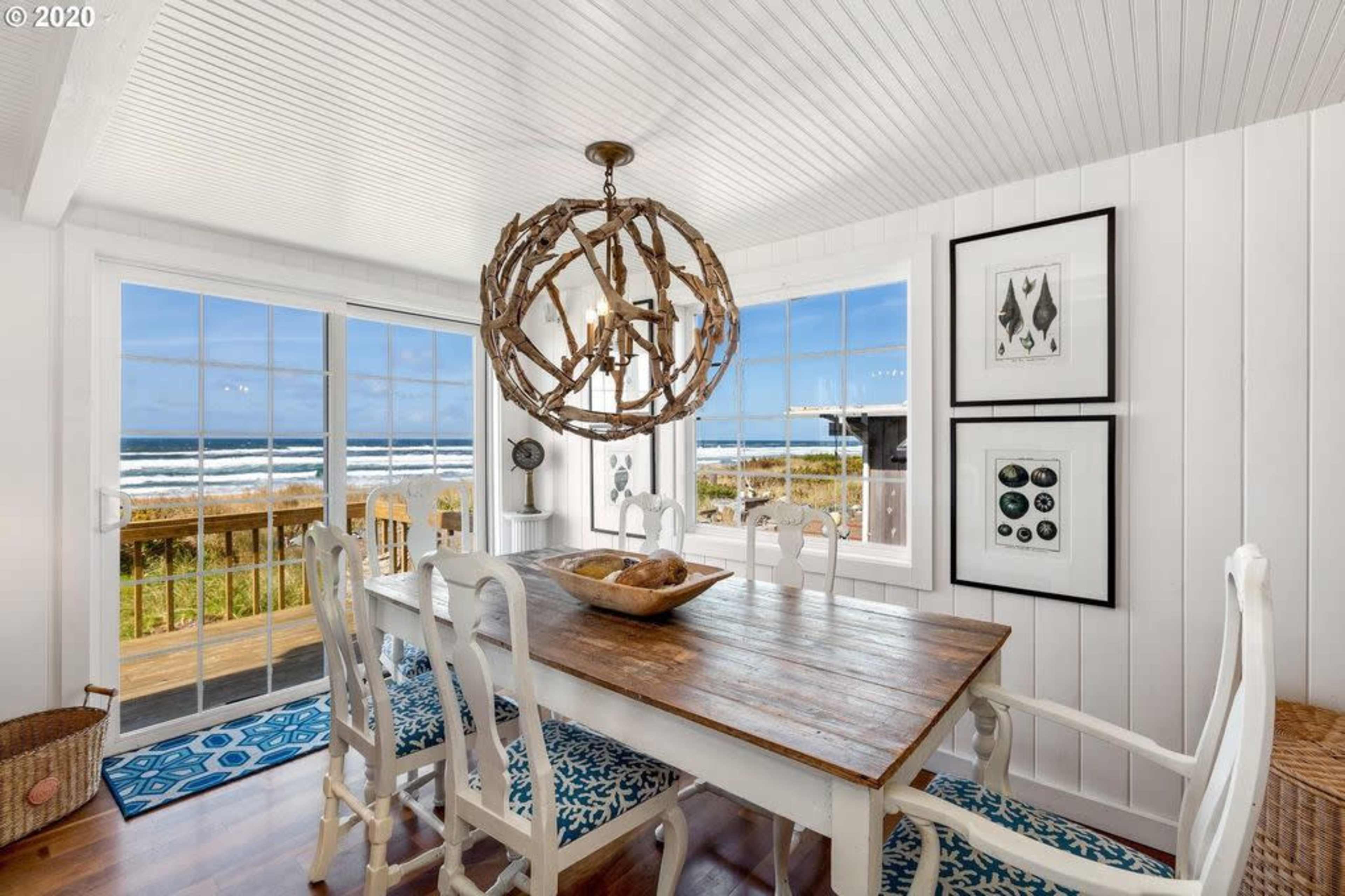 A dining room features a wooden table surrounded by white chairs, with large windows showcasing an ocean view and natural light.