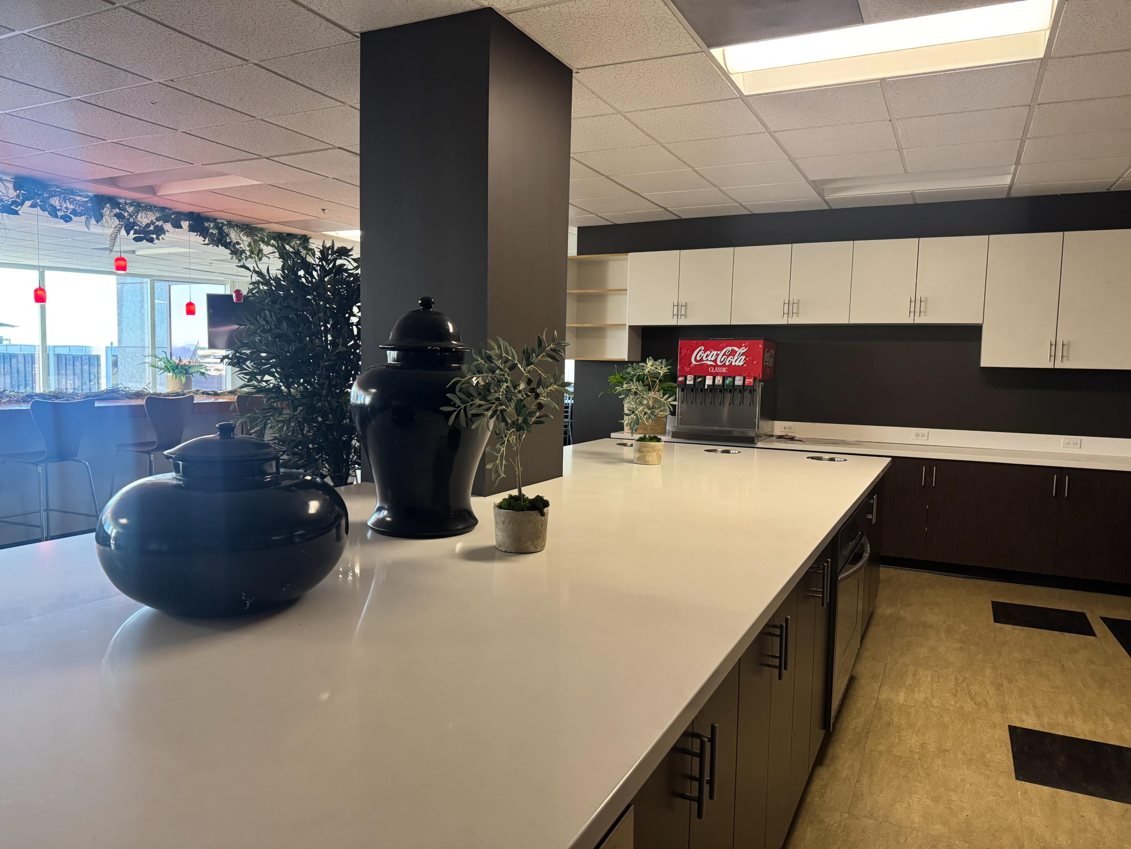 The image shows a modern kitchen area with a large white countertop, black decorative pots, and a Coca-Cola beverage dispenser against a backdrop of cabinets.