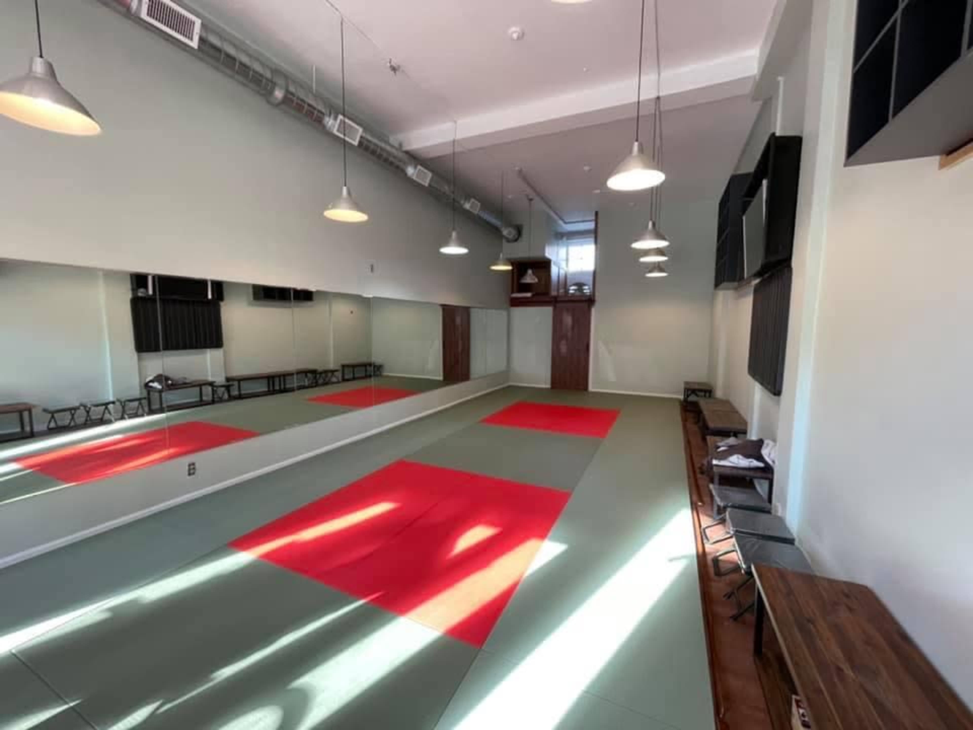 The image shows a martial arts studio with red and gray mats, large mirrors on one wall, and wooden benches along the opposite side.