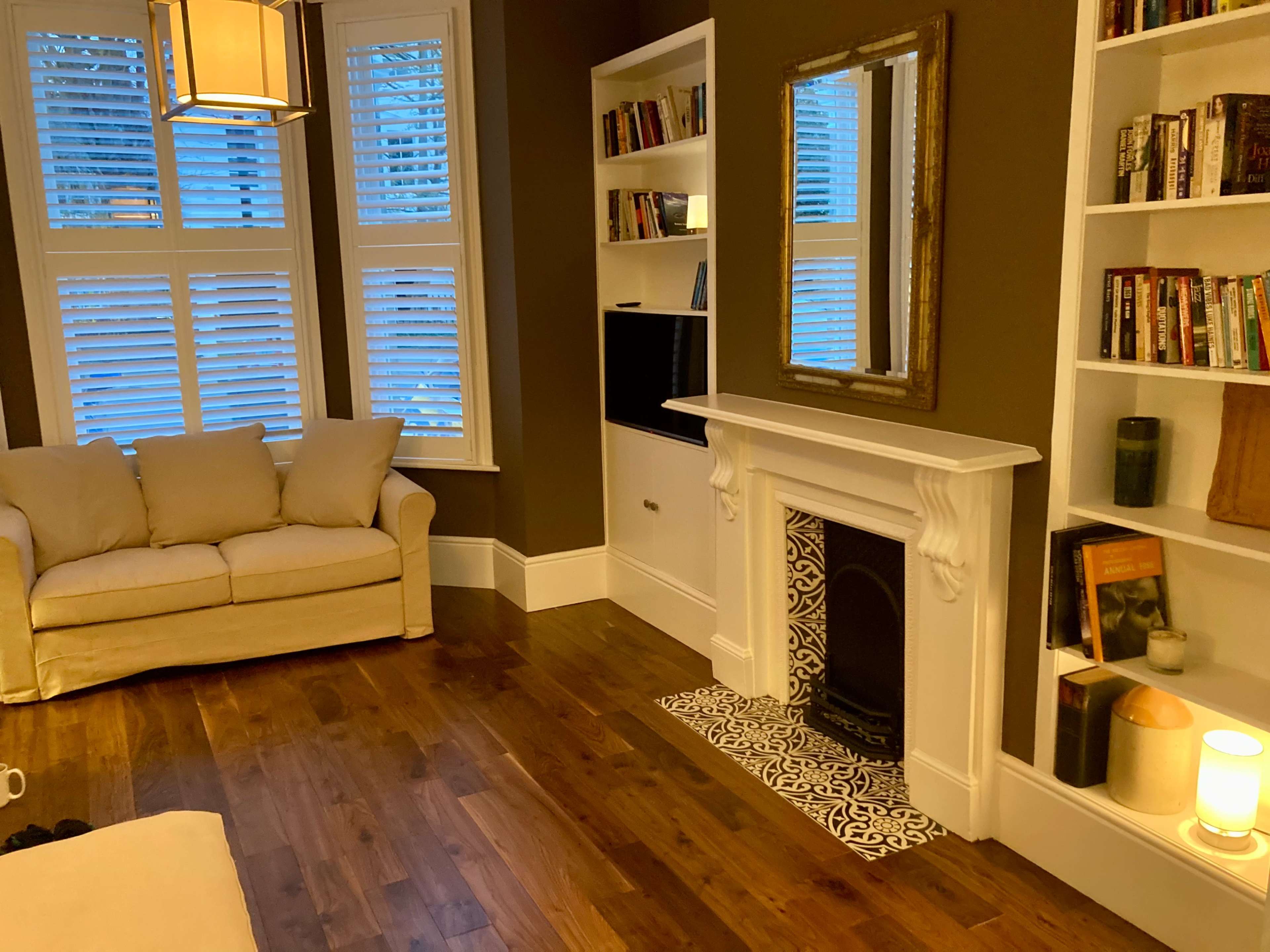 The room features a cream-colored sofa, a flat-screen TV mounted on the wall, a mirrored fireplace with decorative tiles, and bookshelves filled with books, all illuminated by warm lighting.