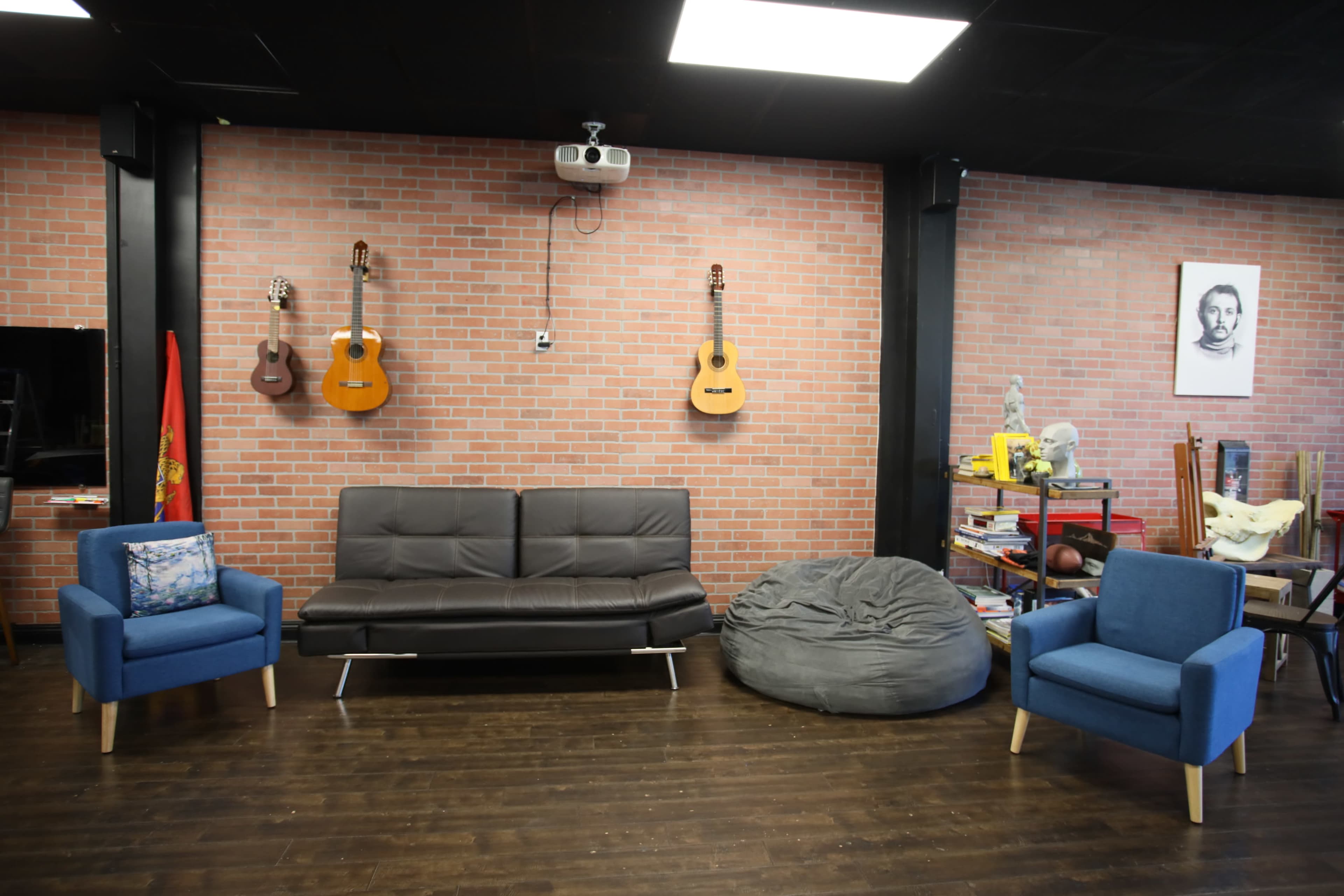 The room features a dark brick wall with acoustic guitars hanging, a grey beanbag chair, a black sofa, and two blue armchairs.
