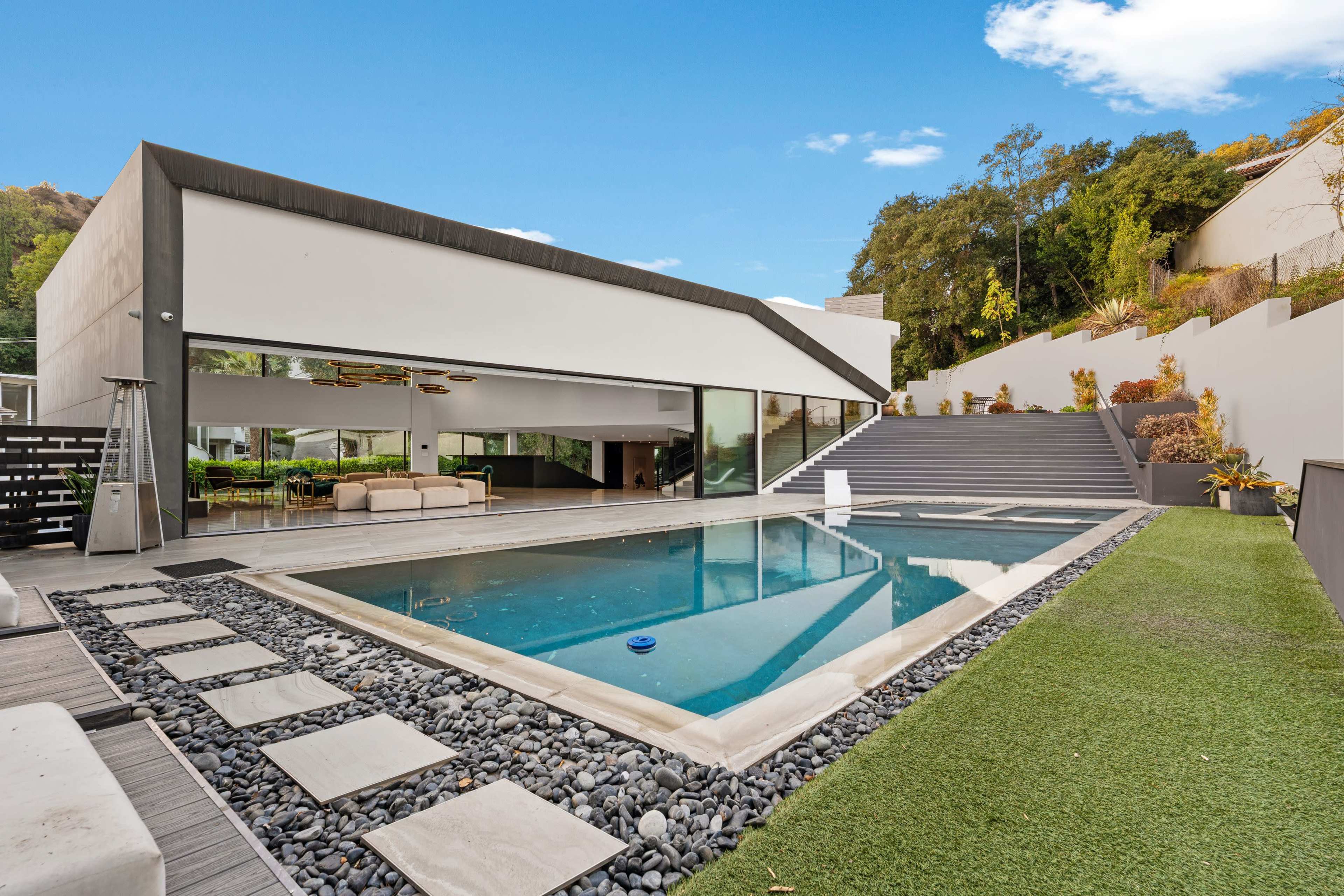The image shows a modern house with a large swimming pool, surrounded by landscaped greenery and stone pathways.