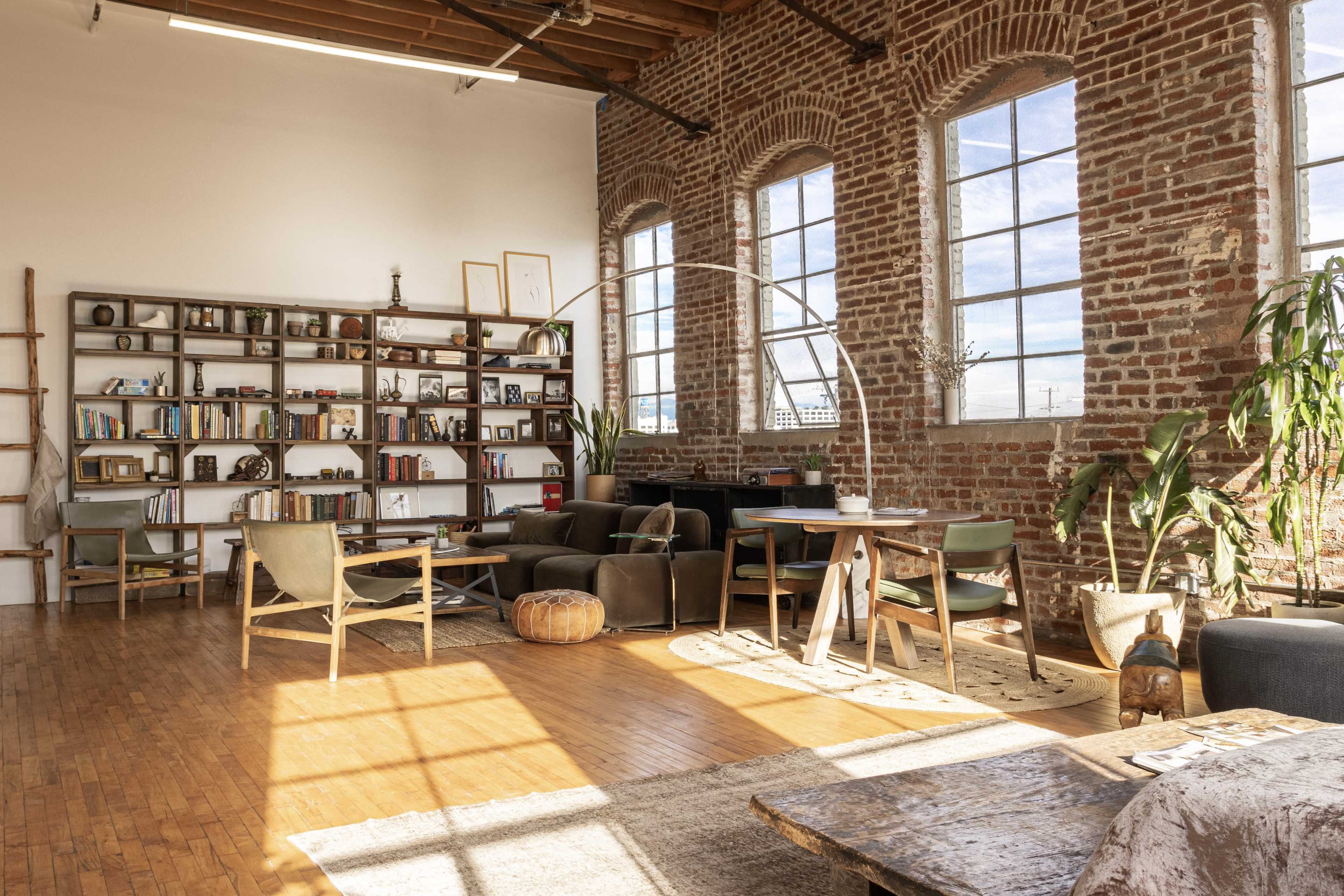 A spacious, sunlit living area with brick walls, a bookshelf filled with books, and comfortable seating arranged around a wooden table.