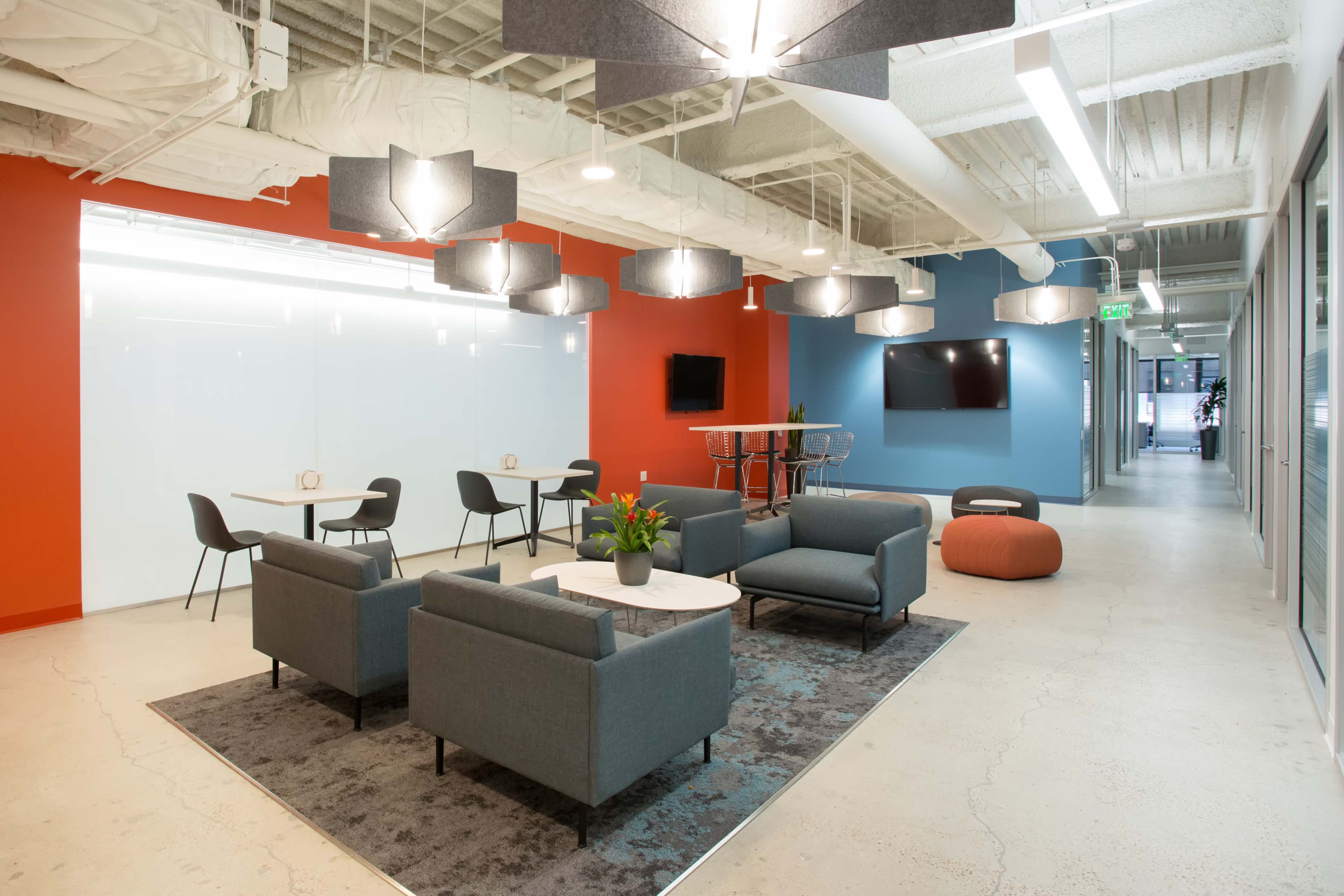 The image shows a modern office space with seating areas, tables, and contemporary lighting fixtures, complemented by blue and red walls.