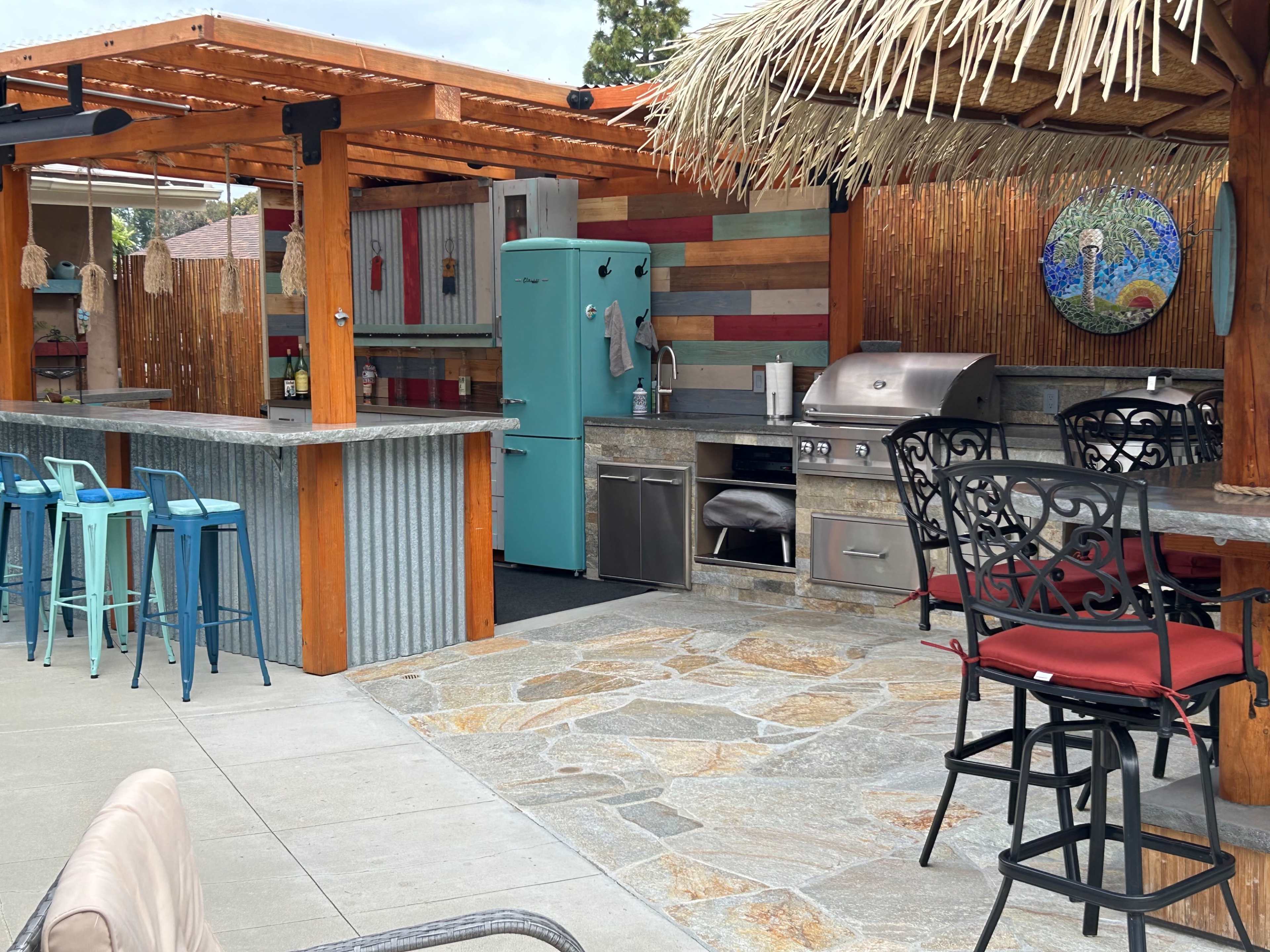 The outdoor kitchen features a stone-paved area with a stainless steel grill, turquoise refrigerator, and bar seating under a thatched roof.