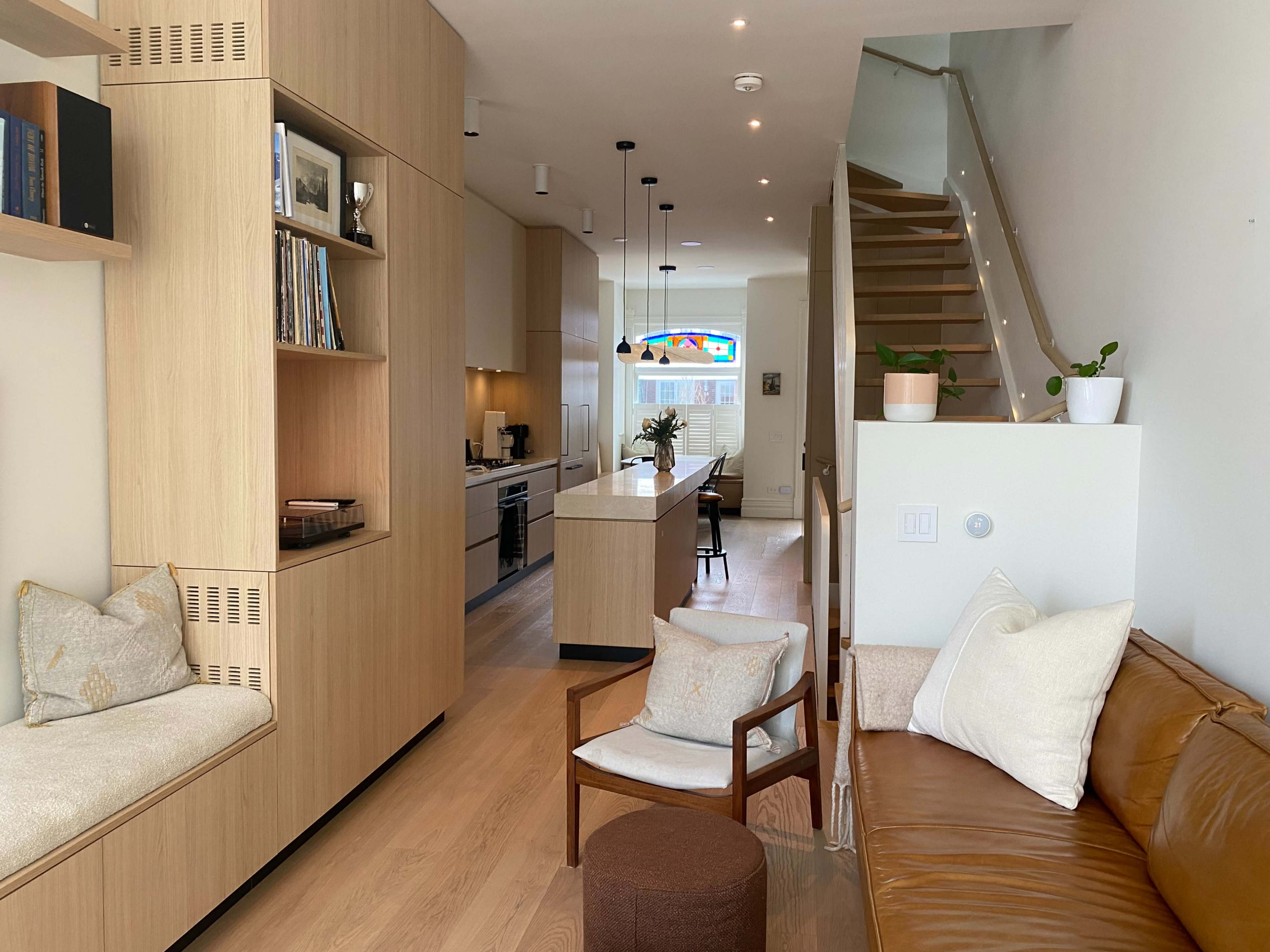 The image shows a modern, open-concept living space featuring a kitchen, dining area, and a staircase, with wooden cabinetry and seating areas.