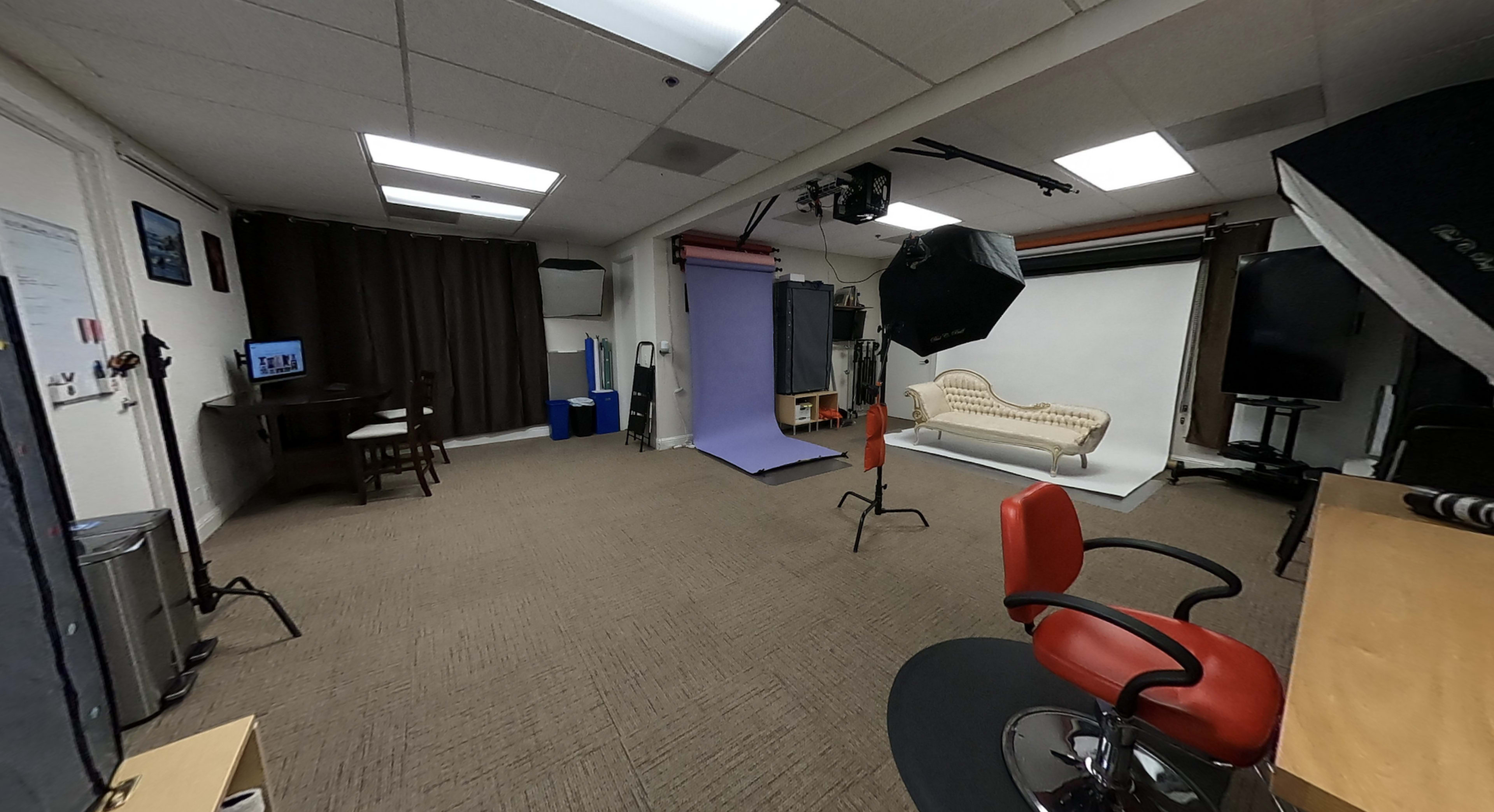 A photography studio featuring a purple backdrop, a white backdrop, a vintage couch, and various lighting equipment arranged in a well-lit room.
