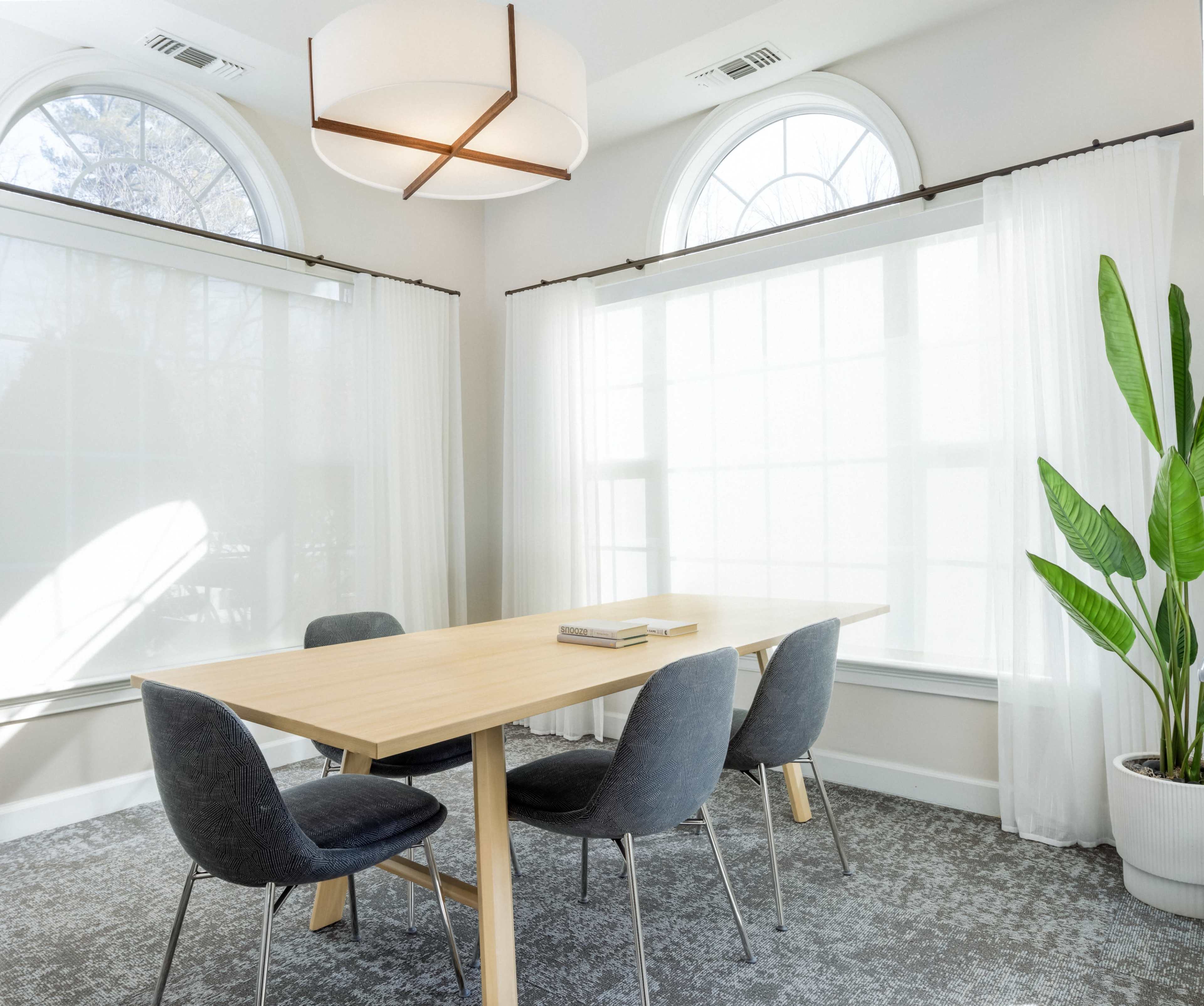 A wooden dining table is surrounded by four gray chairs in a brightly lit room with large windows and sheer curtains.