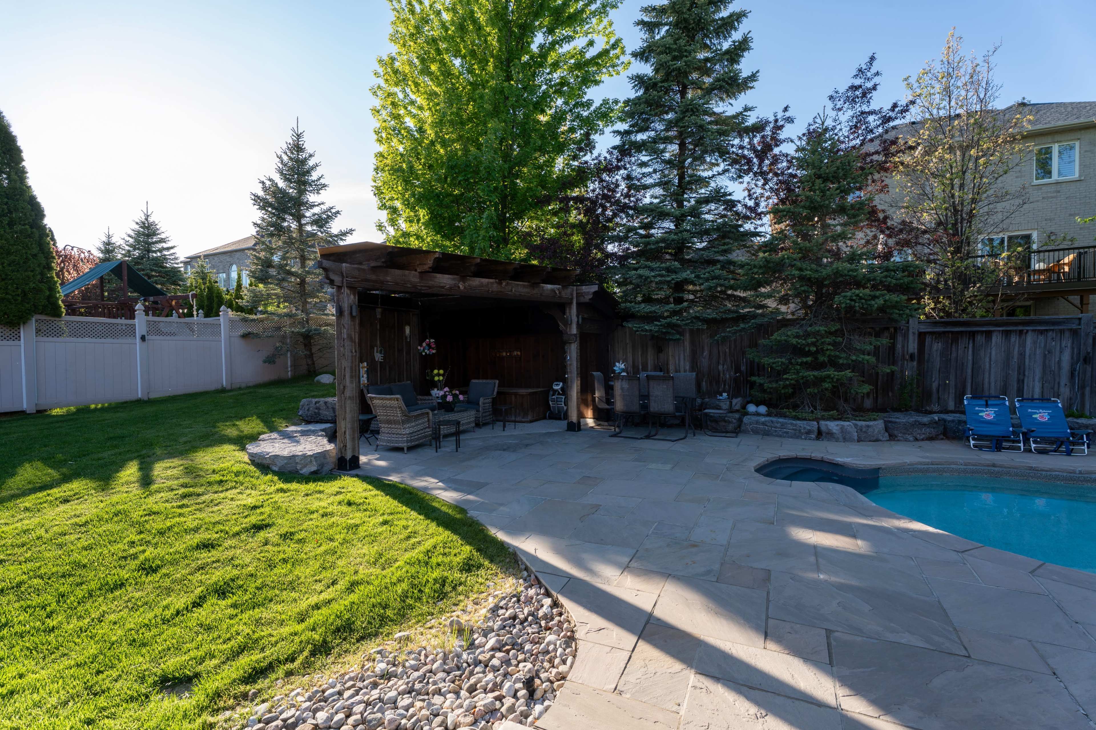 The image shows a landscaped backyard with a stone patio, a wooden gazebo, and a swimming pool, surrounded by trees and a fence.