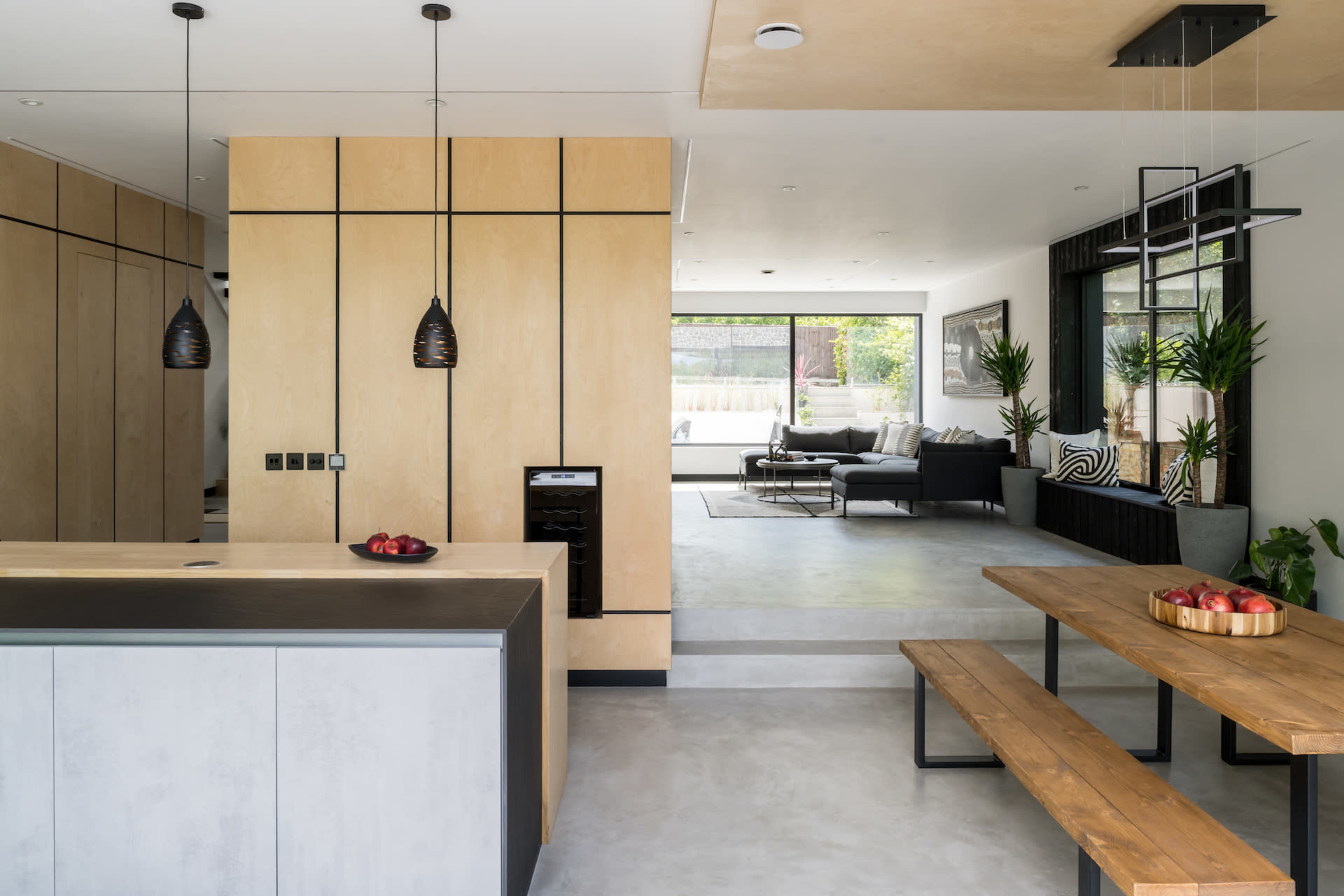 A modern, open-concept living space featuring a kitchen with light wood cabinetry, a dining area with a wooden table, and a lounge with large windows overlooking a garden.
