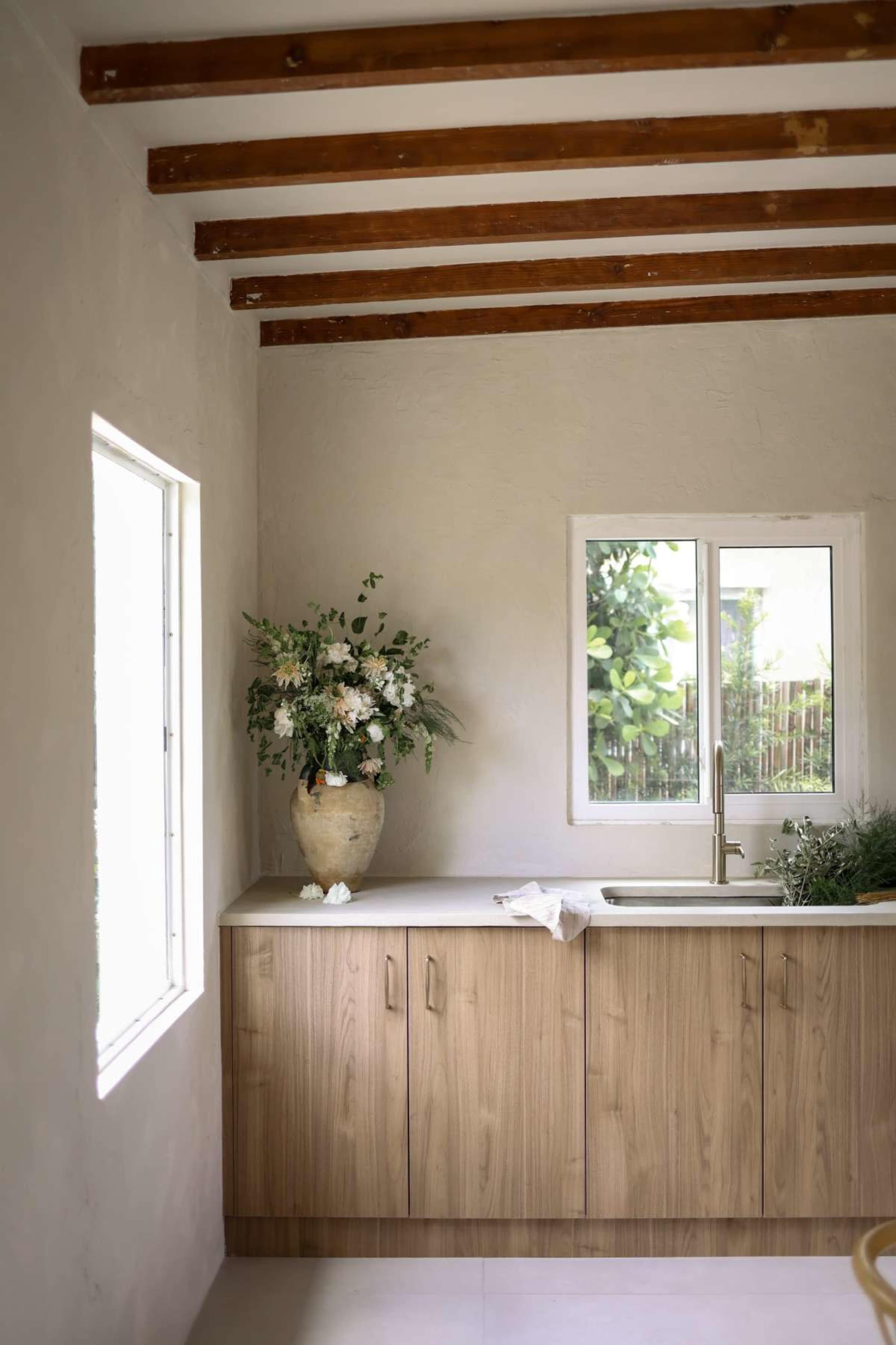 A kitchen with wooden beams on the ceiling features a light-colored cabinetry, a sink, and a large vase with greenery near the window.