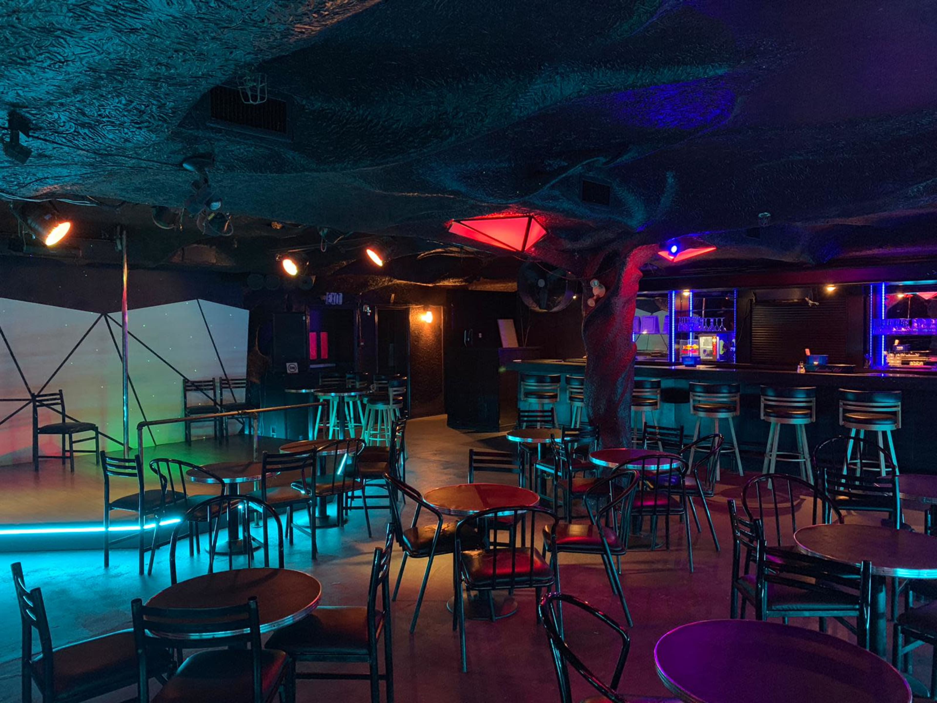 The image shows a dimly lit nightclub interior featuring circular tables and a bar area illuminated with colorful lights.