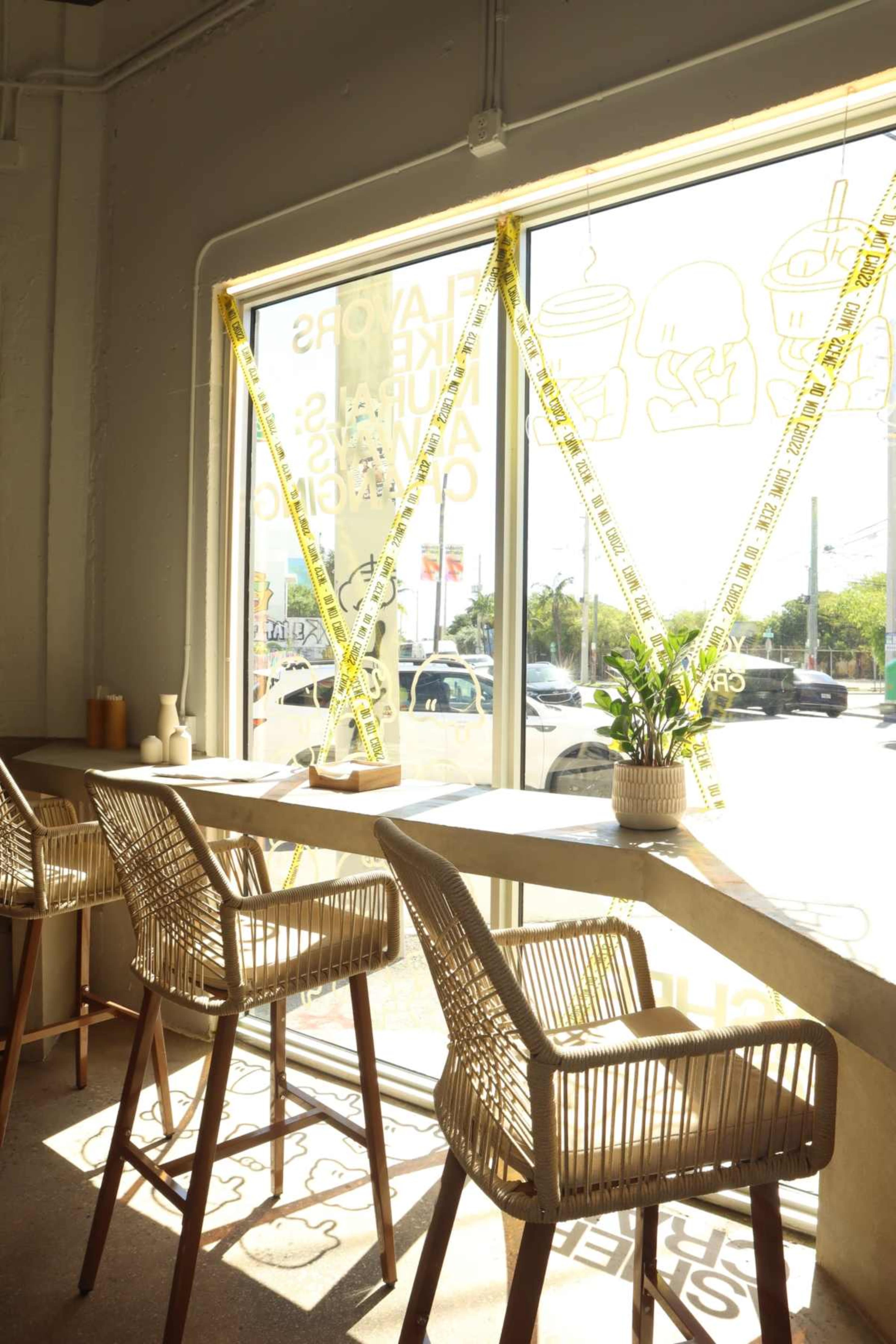 The image shows a bright interior of a café with rattan barstools arranged by a large window, featuring yellow tape with graphics and a small plant on the counter.
