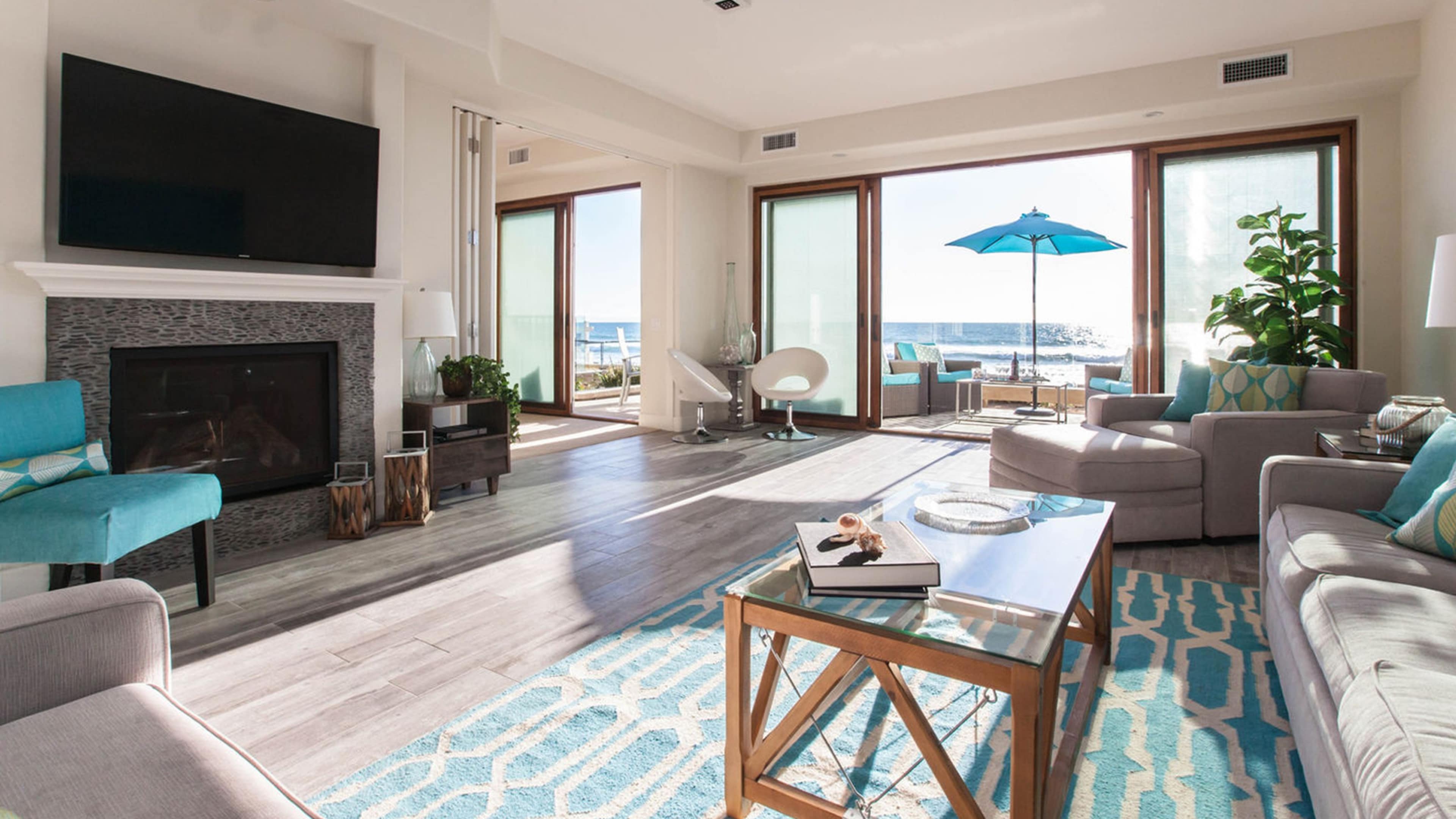A modern living room with large windows facing the ocean, featuring a fireplace, comfortable seating, and a coffee table on a patterned rug.