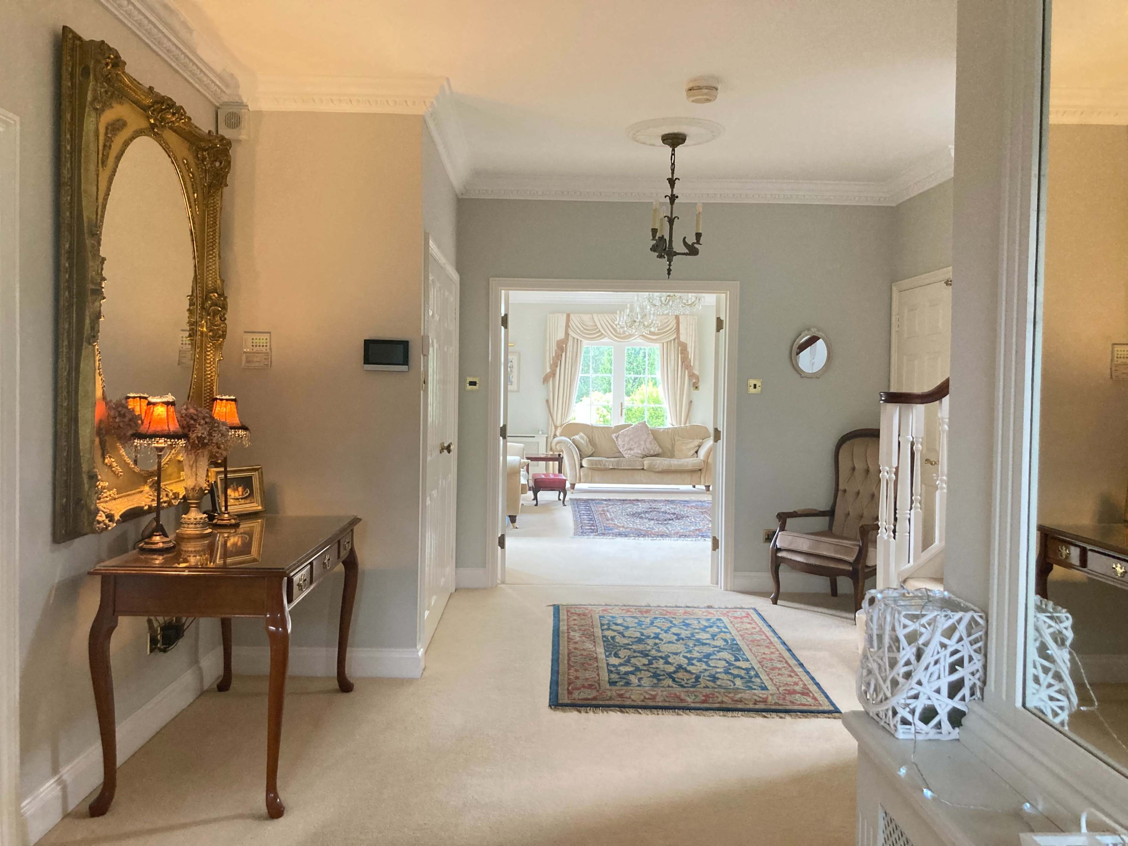 A well-lit hallway featuring a large ornate mirror, a wooden console table with lamps, and an open doorway leading to a living room adorned with a chandelier and upholstered furniture.