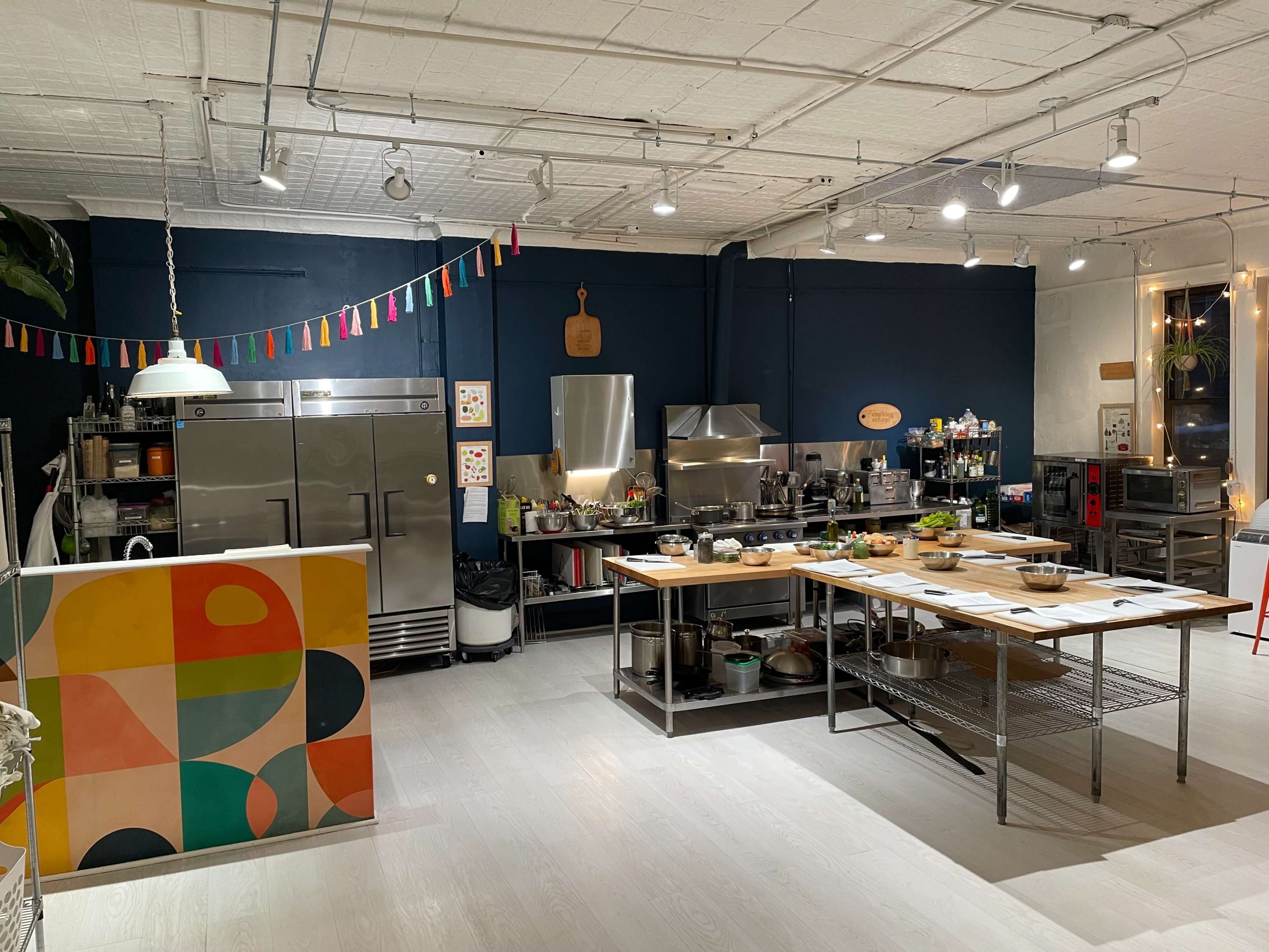The image shows a modern kitchen with stainless steel appliances, a large central table, and colorful decorations on the walls.