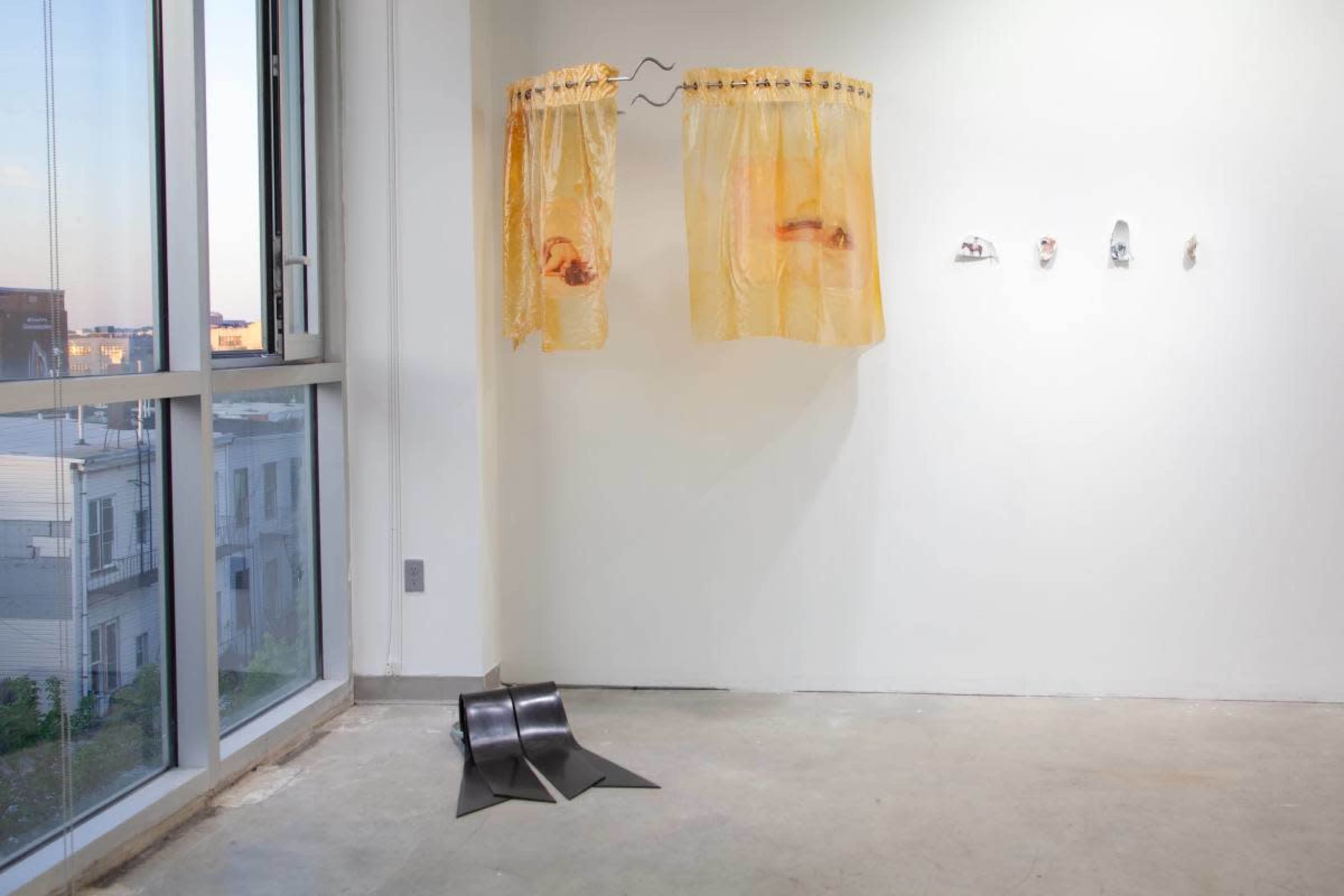 The image shows an art gallery interior featuring yellow sheer curtains and several small sculptures mounted on a white wall, with a curved metal sculpture on the floor.