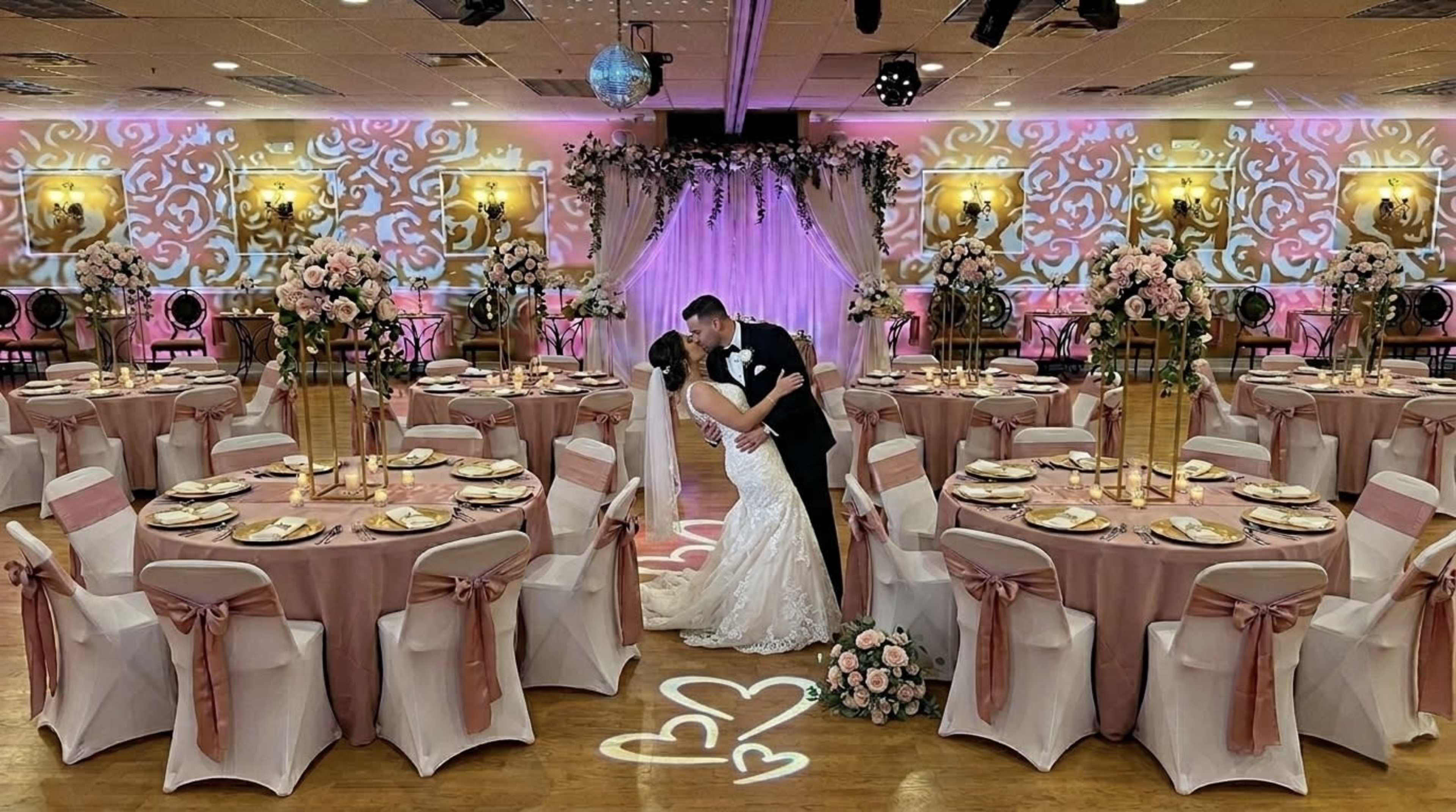 A newlywed couple shares a kiss in a decorated reception hall with elegantly set tables and floral arrangements.