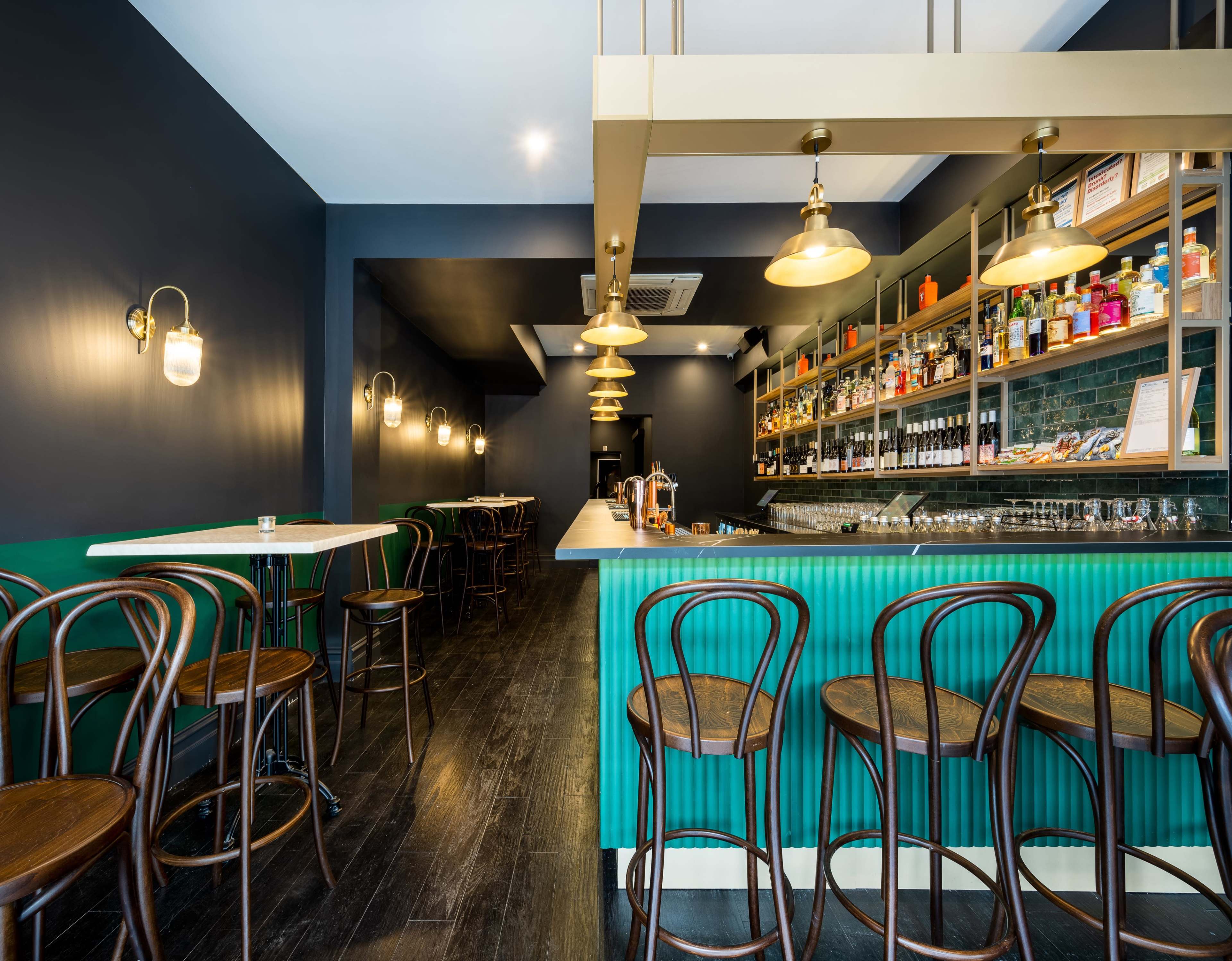 The interior of a modern bar features sleek green and gold decor, with a long counter and brown wooden stools against a dark wall.