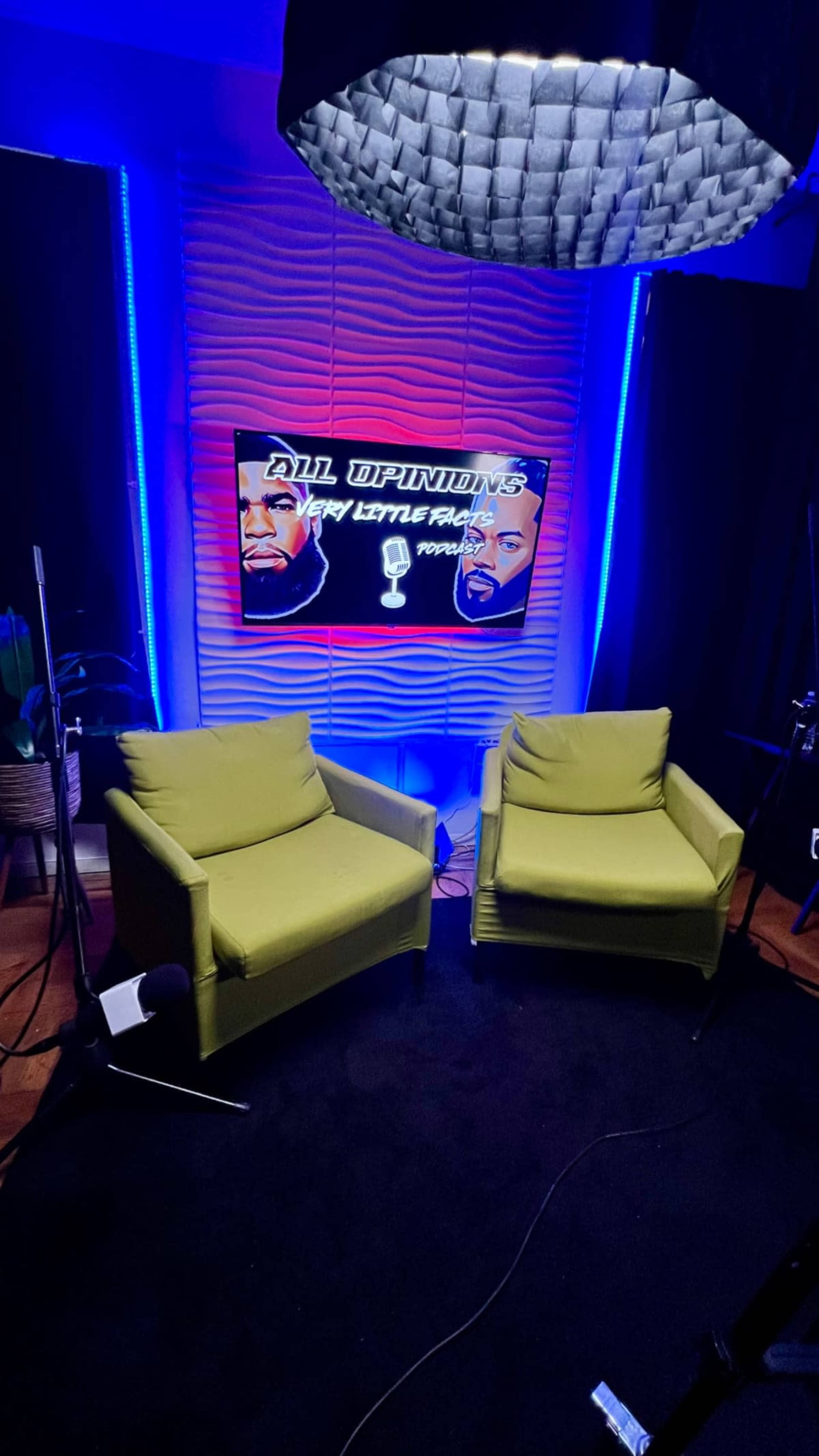 The image shows a brightly lit studio setup featuring two green chairs facing a screen displaying the text "All Opinions" with graphics of two faces in the background.
