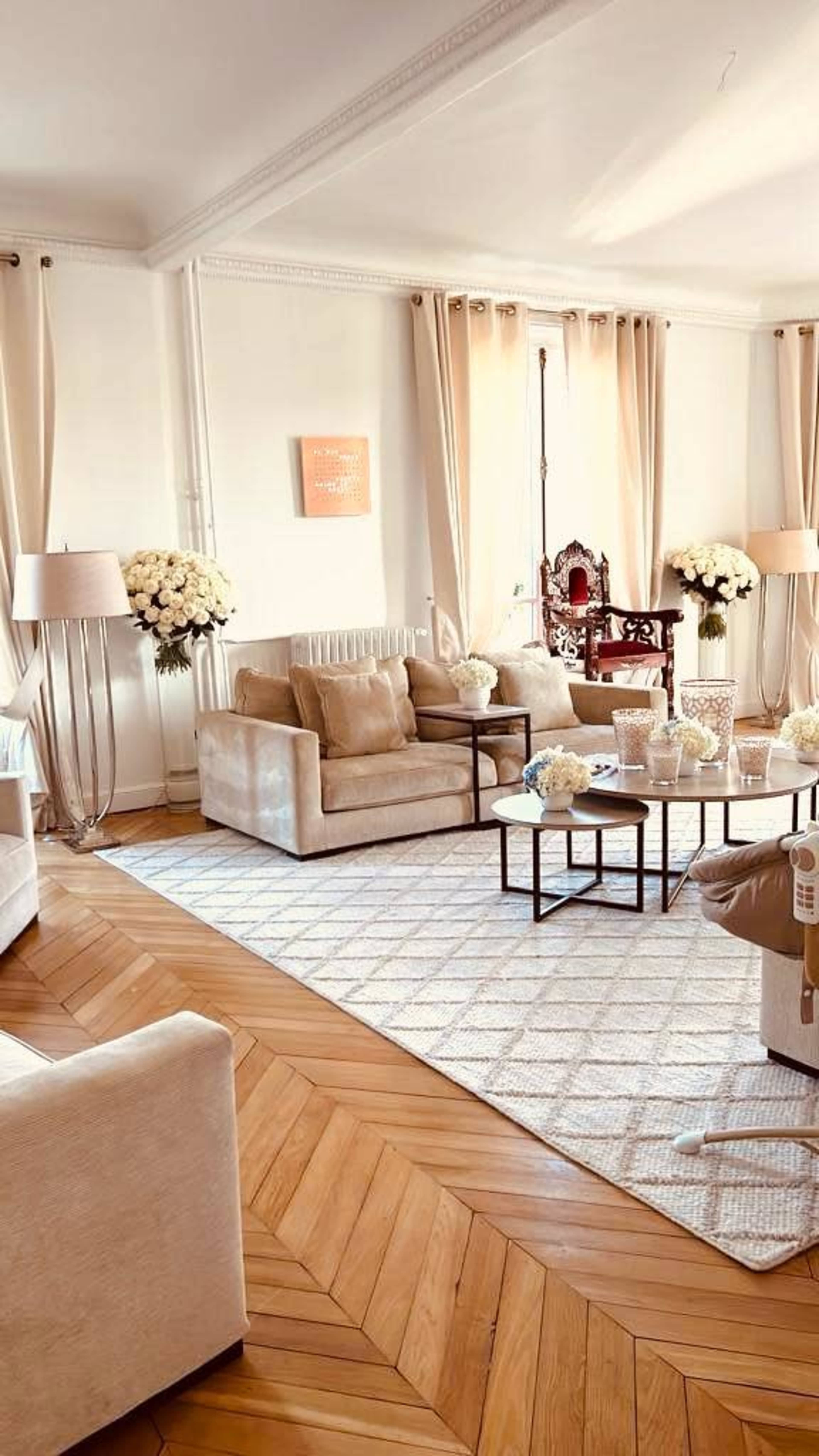 The living room features two beige sofas and a coffee table, surrounded by floral arrangements and large windows draped with light curtains.