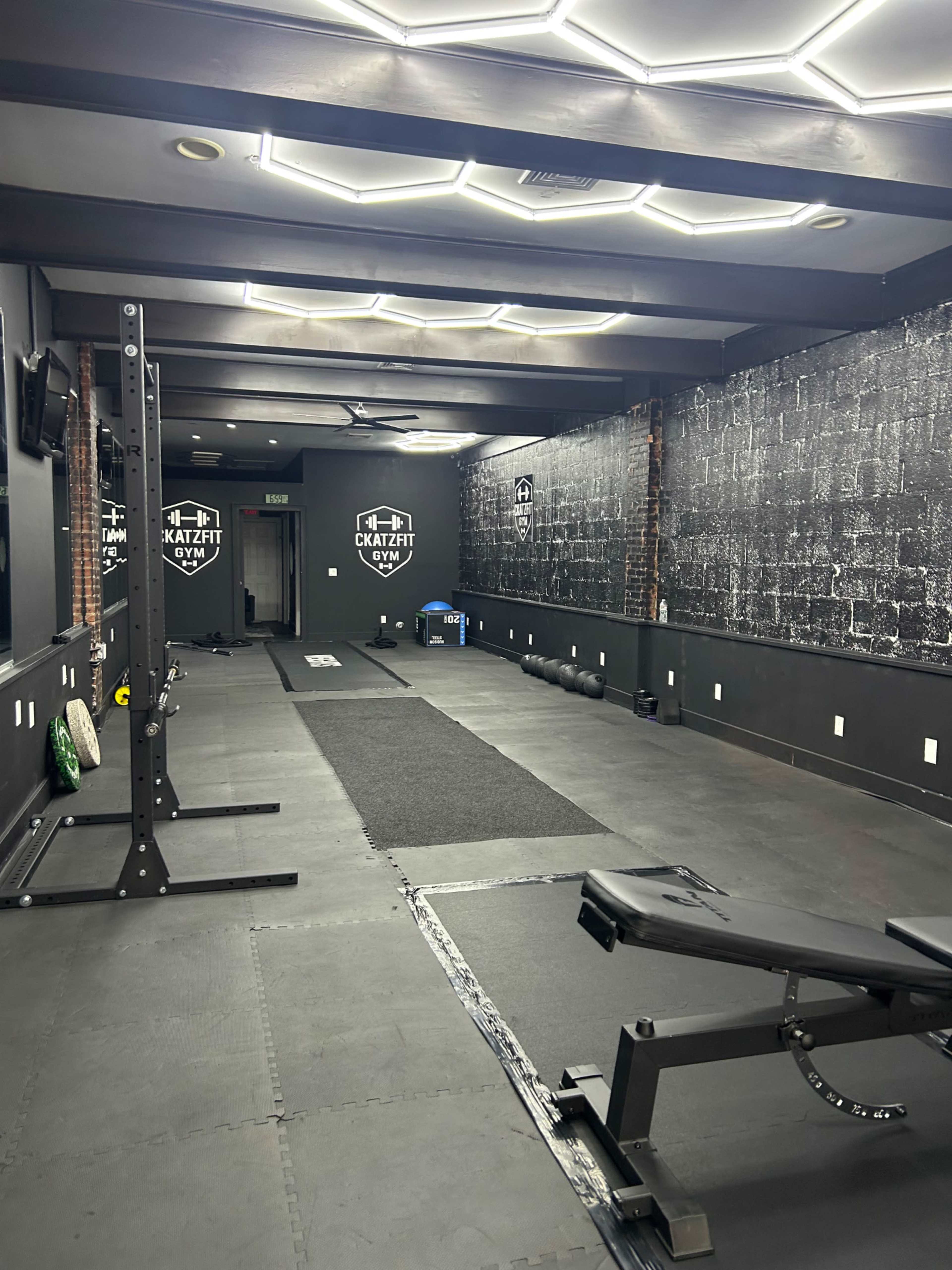 The image shows a black-walled gym interior with workout equipment, including a bench, weights, and exercise mats, under bright overhead lighting.