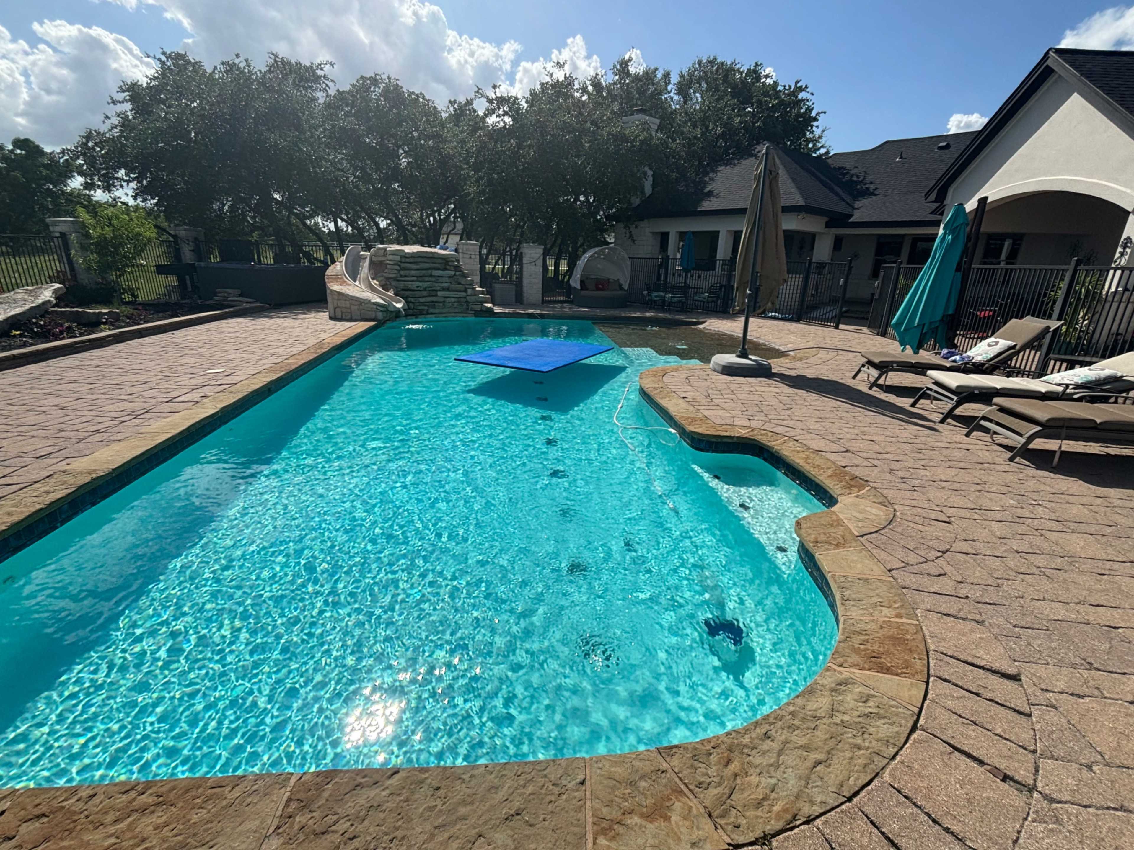 The image shows a clear swimming pool with a blue slide and lounge chairs on the surrounding deck, framed by trees and a house in the background.