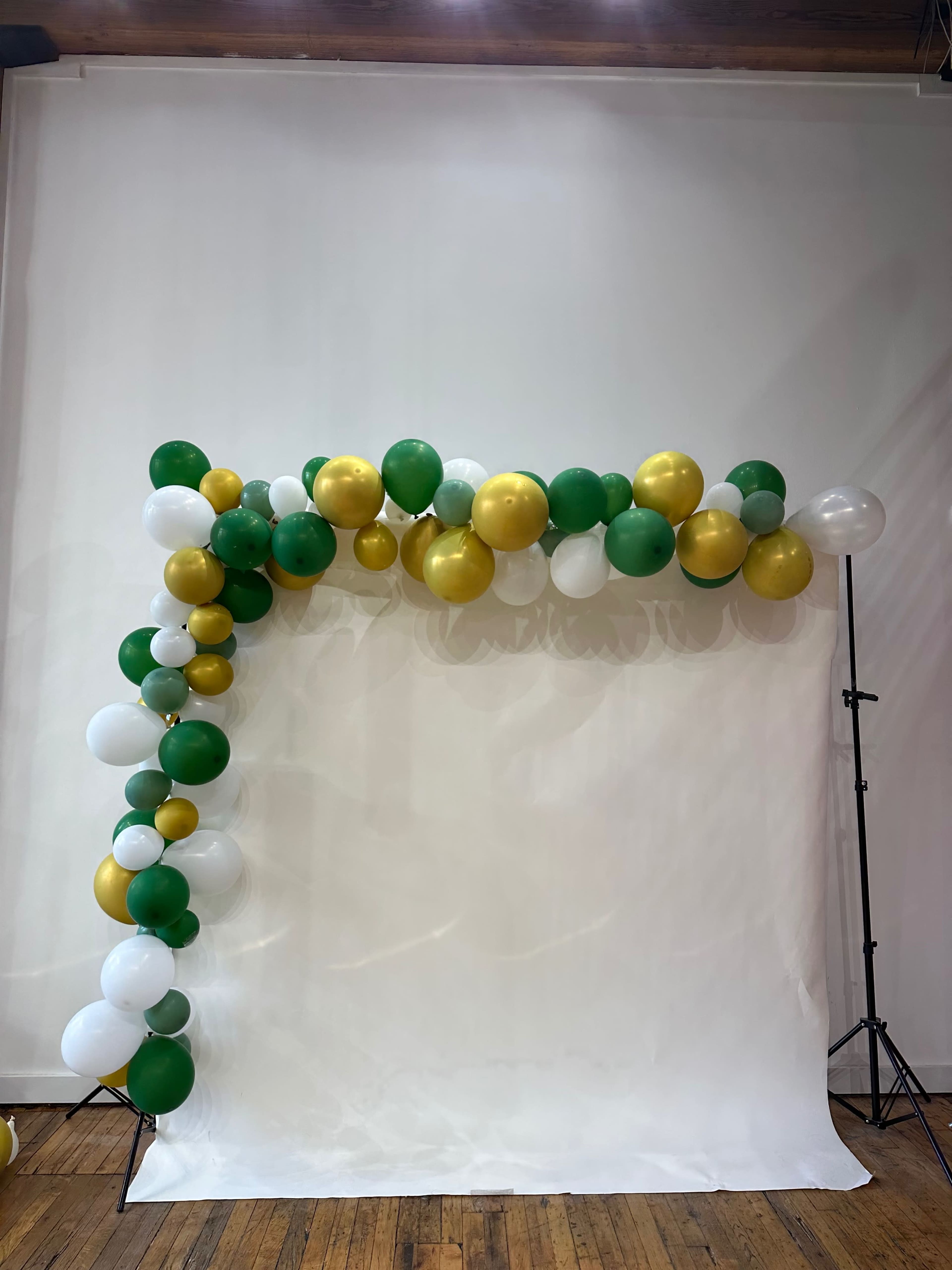 A balloon arch made of green, white, and gold balloons is arranged against a plain white backdrop on a wooden floor.