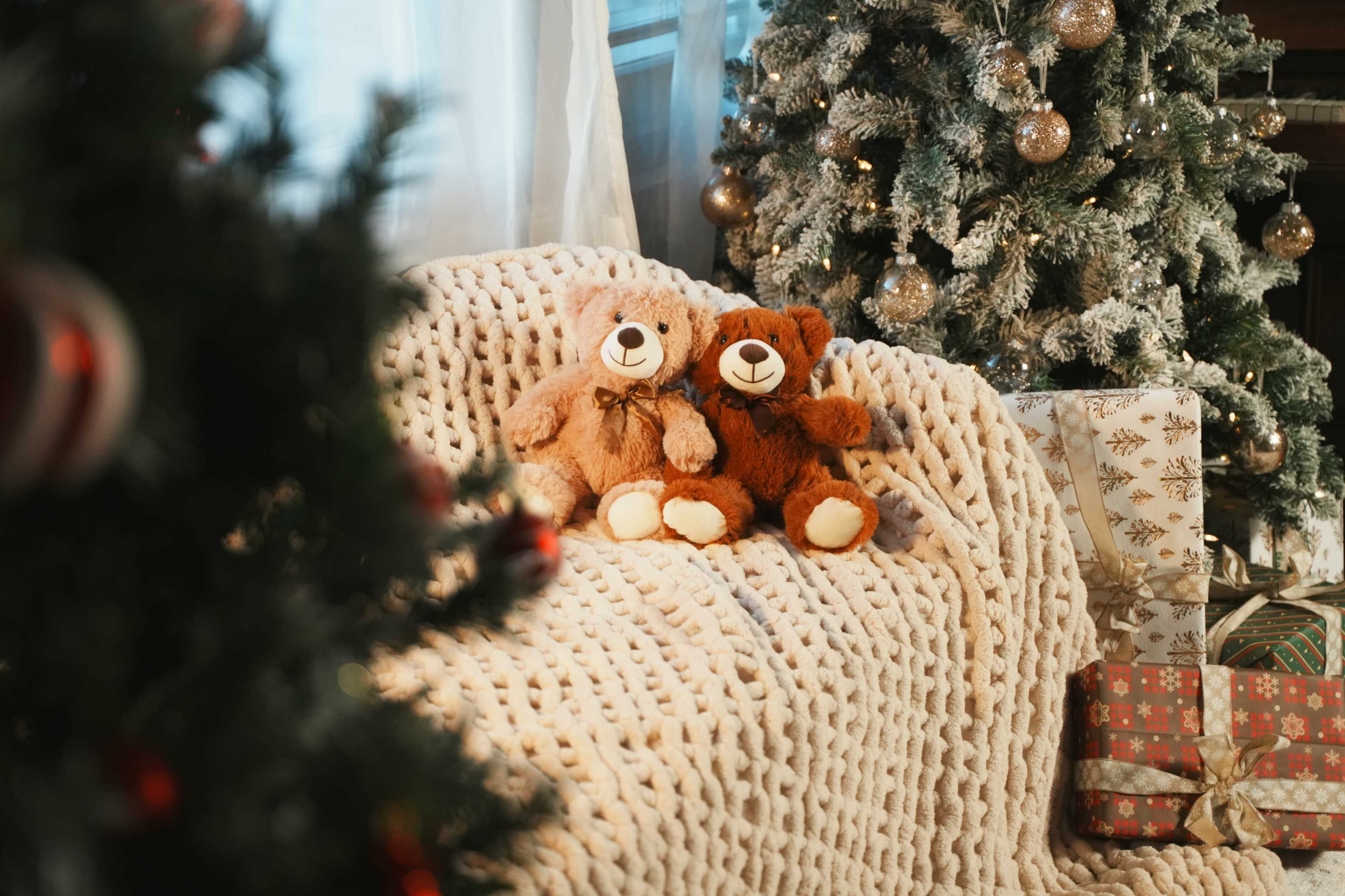Two teddy bears sit on a knitted blanket in front of a decorated Christmas tree and wrapped gifts.