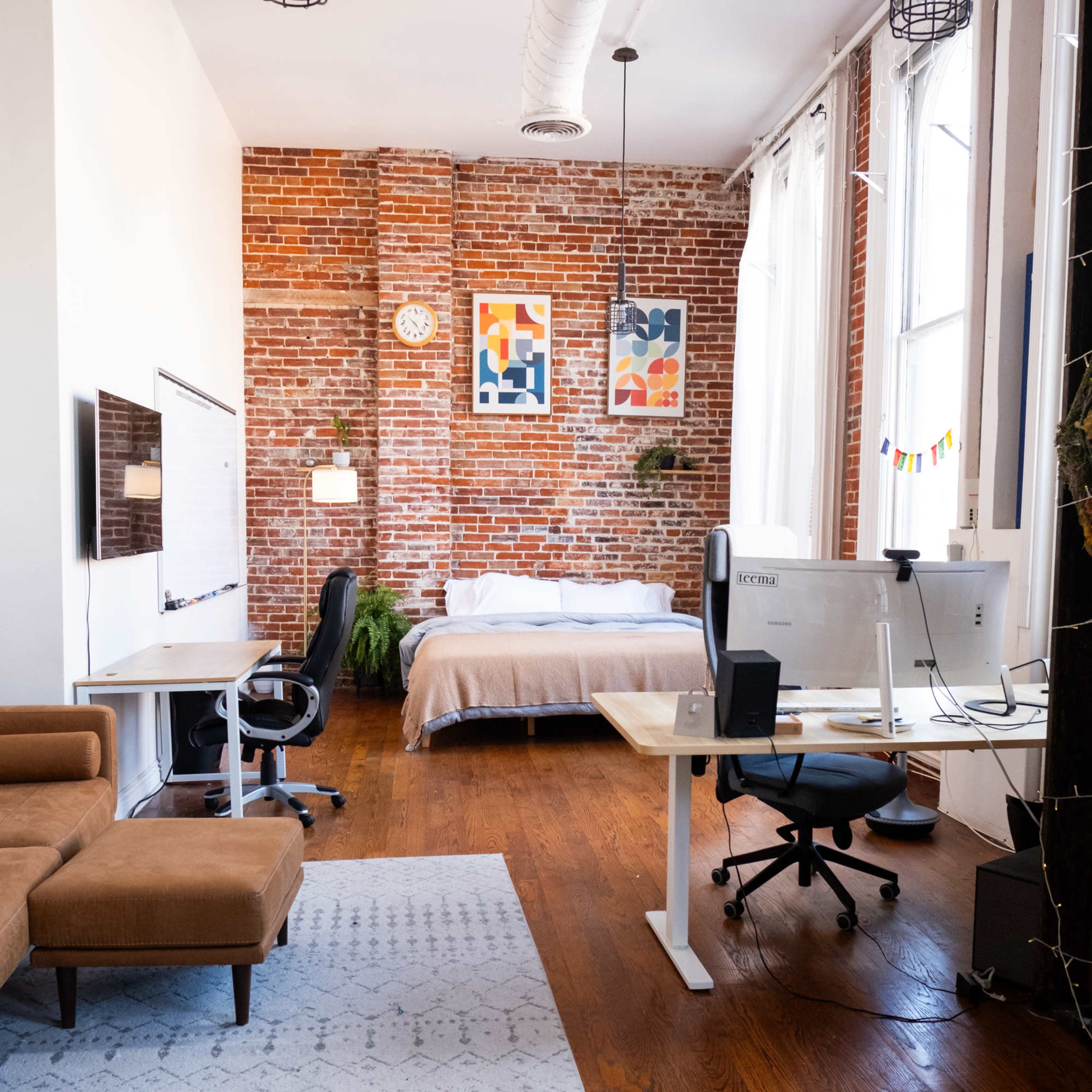 The image shows a spacious studio apartment featuring exposed brick walls, a bed, a workspace with a desk and computer, and comfortable seating.