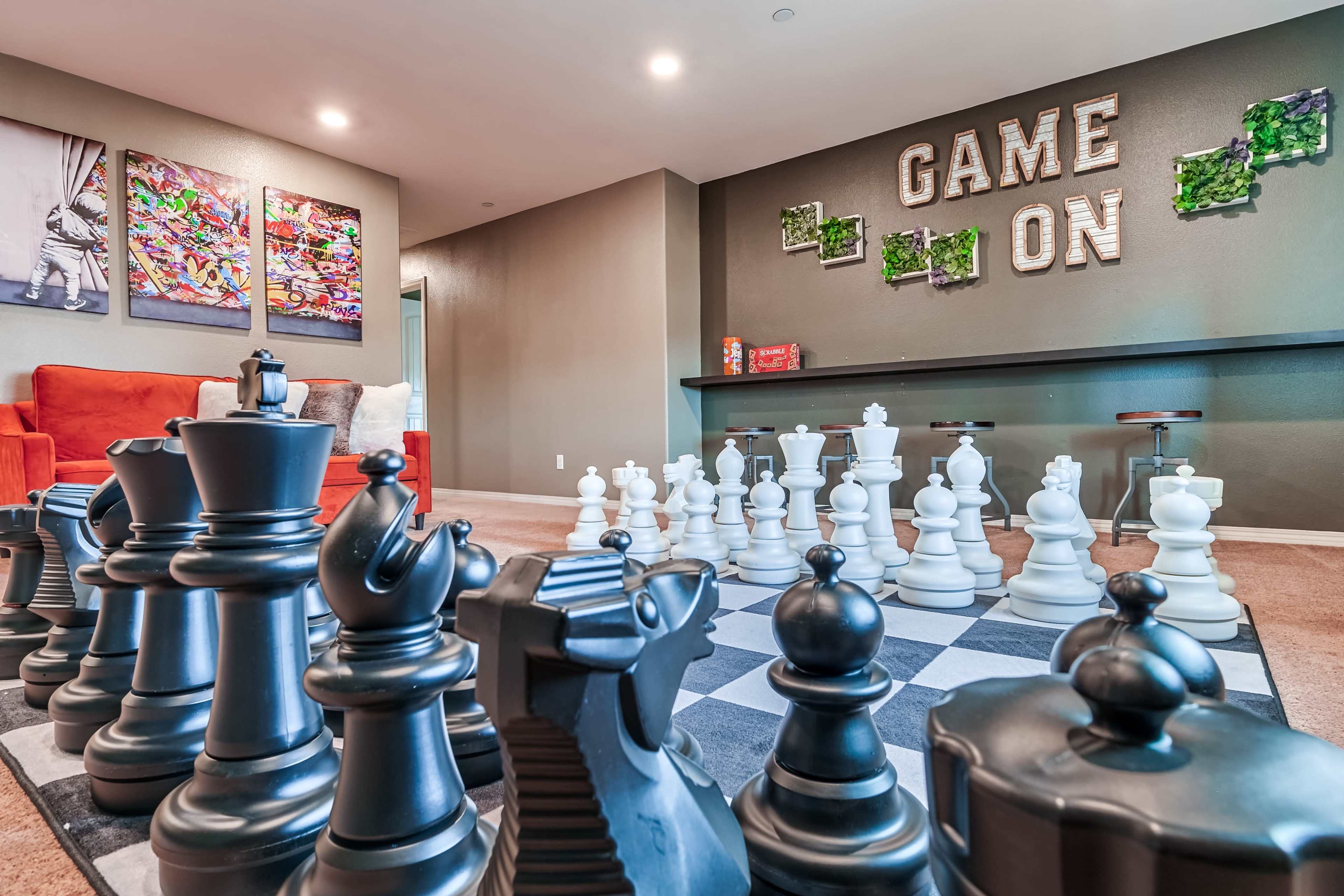The image features a large chess set made of black and white pieces arranged on a checkered board in a game room decorated with colorful artwork and the words "GAME ON" on the wall.
