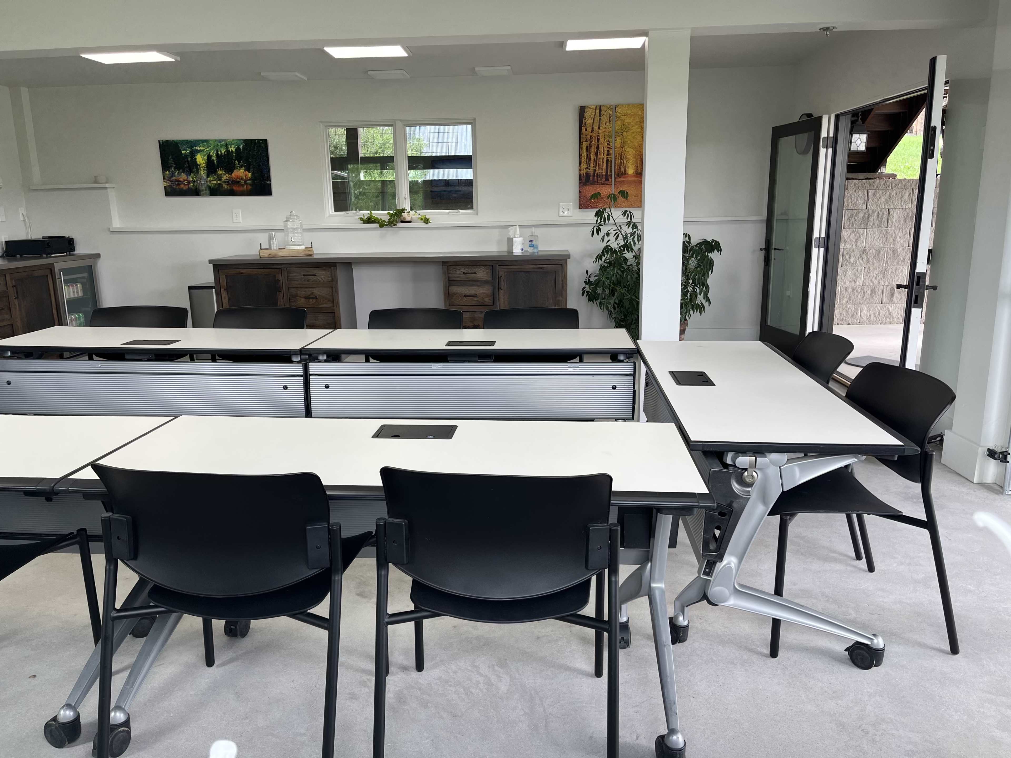 A modern, minimalist classroom setup with black chairs and modular tables arranged for group work, complemented by a console and plants in the background.