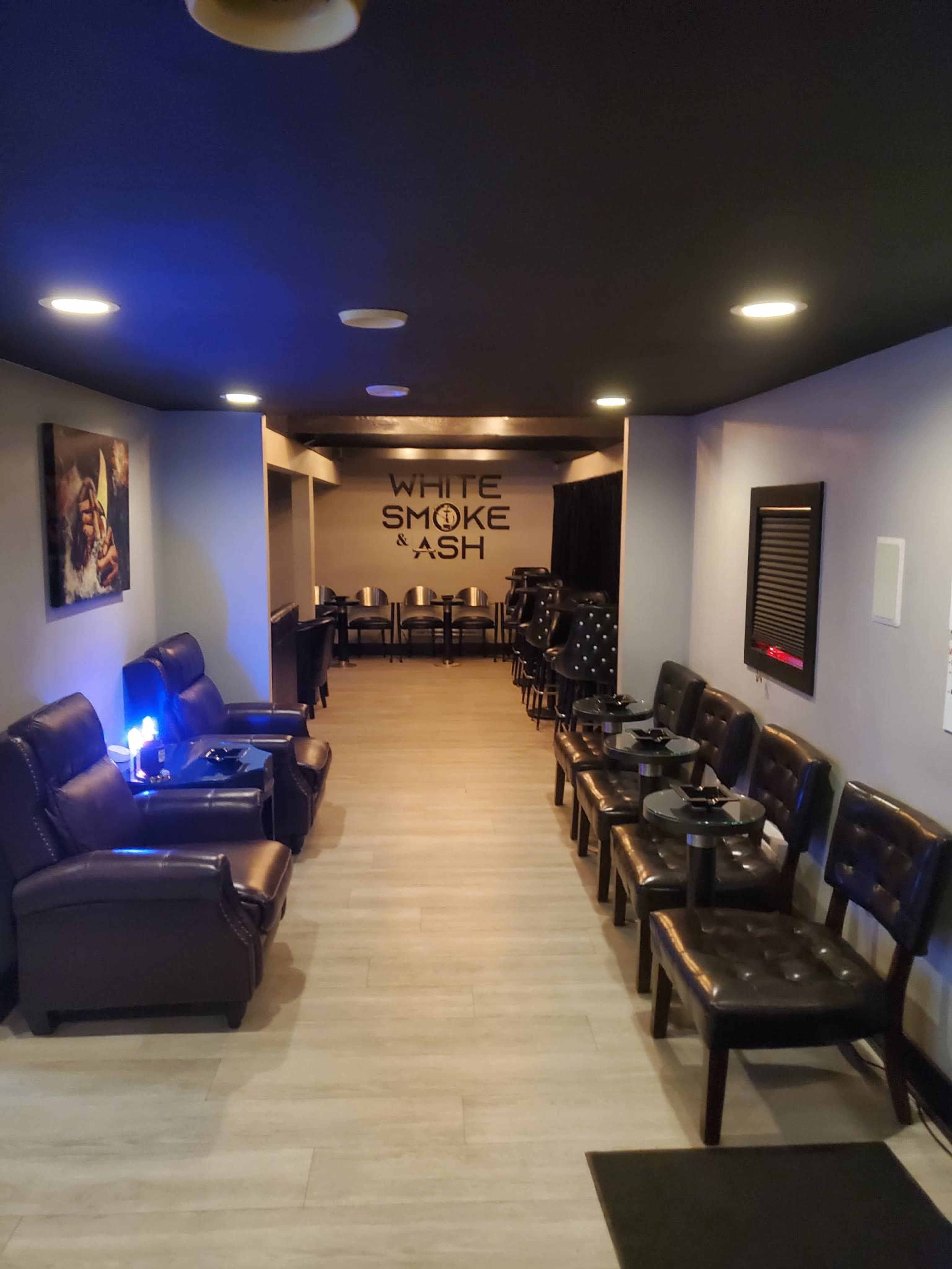 The image shows a narrow hallway leading to a dining area, featuring brown leather chairs and a wall with the text "WHITE SMOKE & ASH."