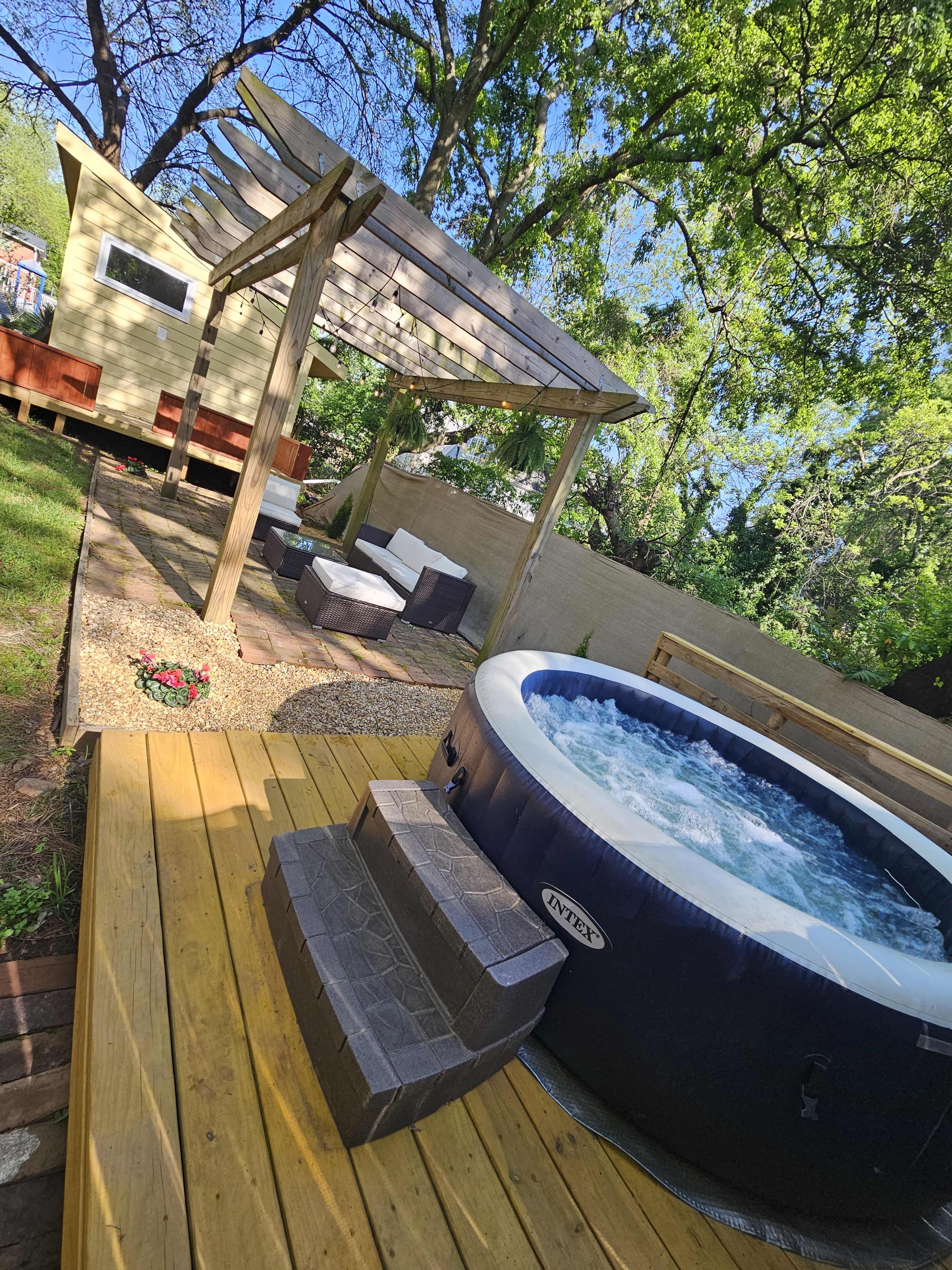 The image shows a backyard scene featuring a hot tub next to a wooden platform and a covered seating area surrounded by greenery.