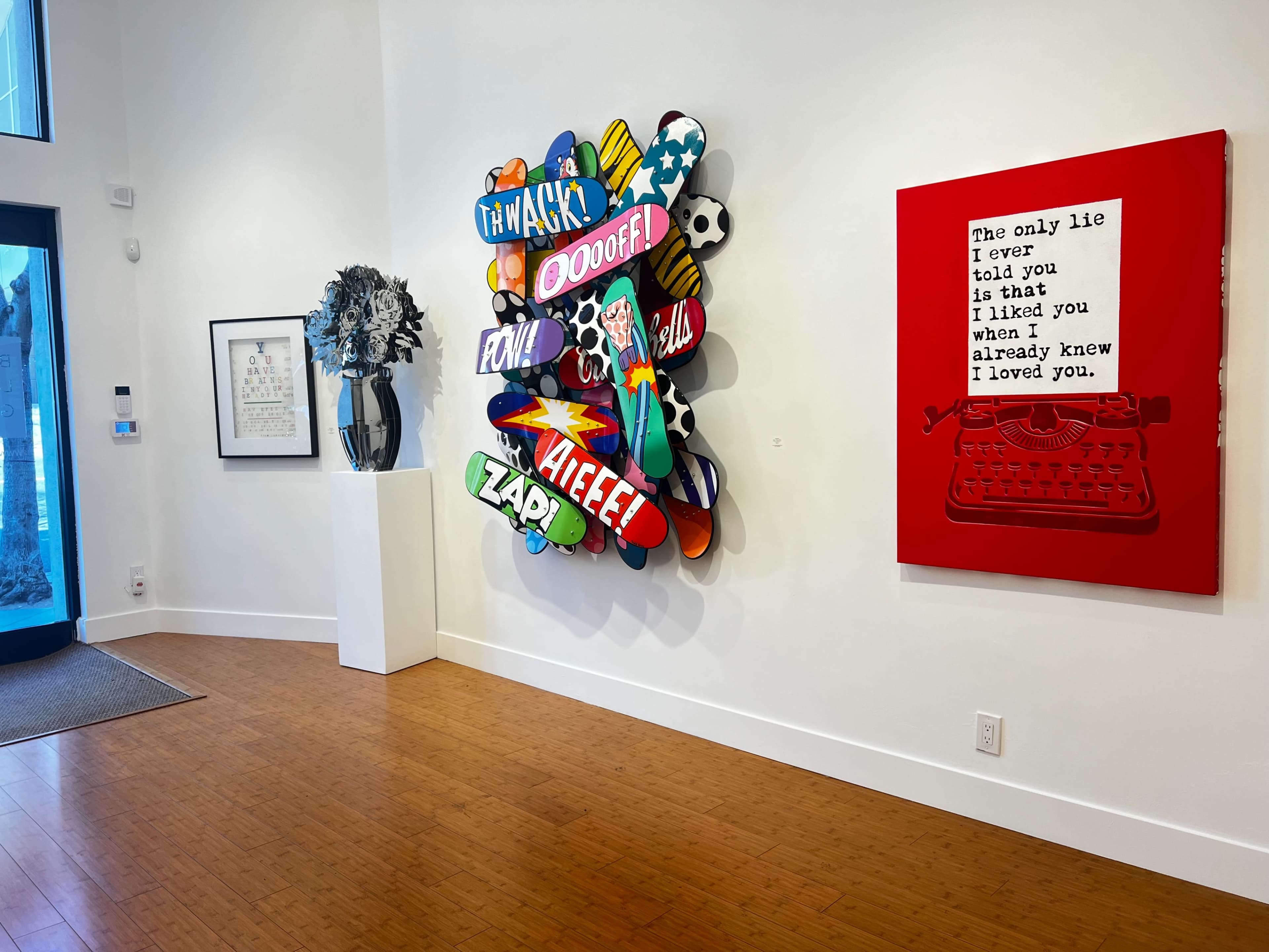 The image shows an art gallery featuring a colorful wall sculpture with comic-style graphics on one side and a red canvas with text on the other, alongside a framed artwork.